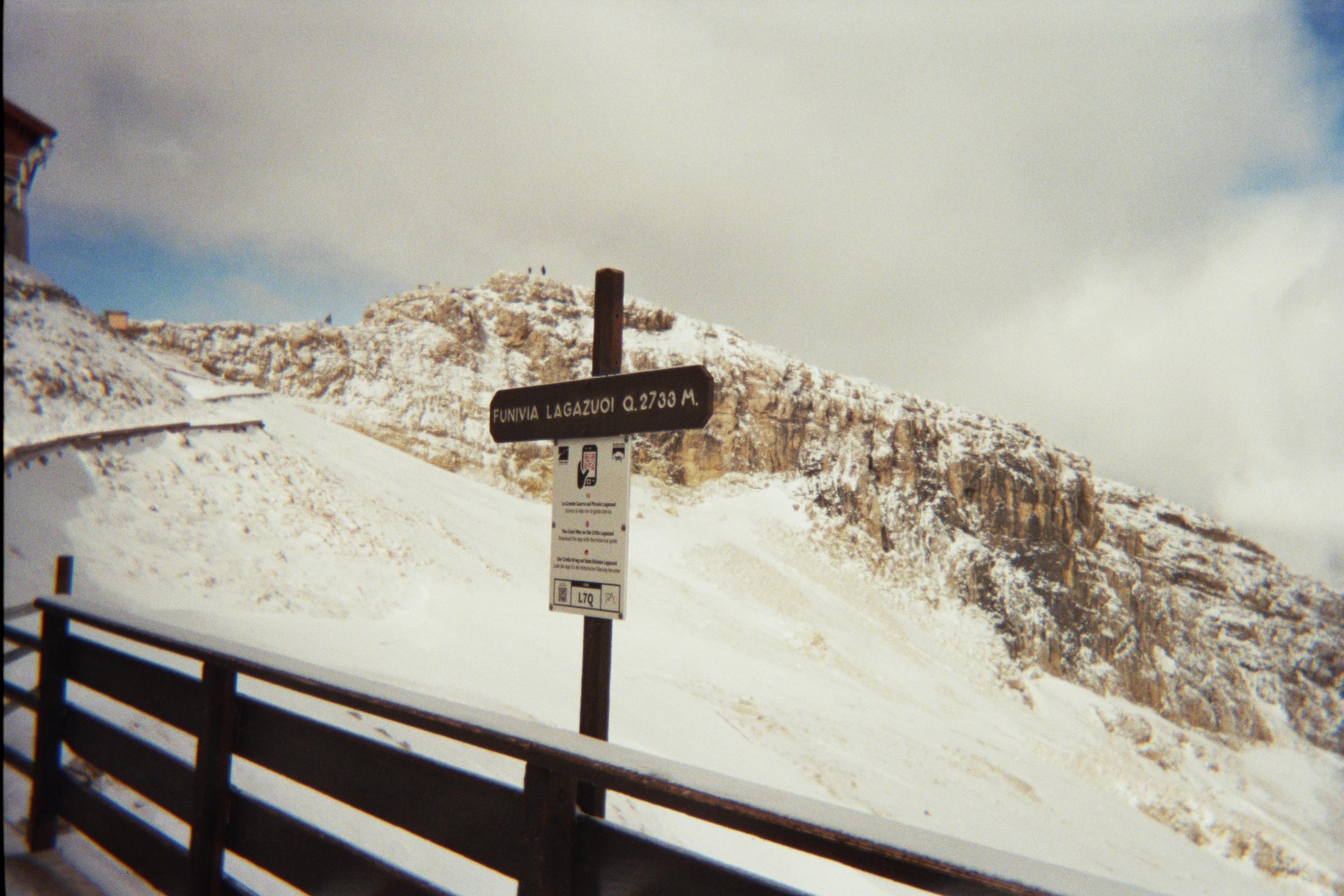 Snow-covered mountain path with a brown wayfinding sign above a metal railing.