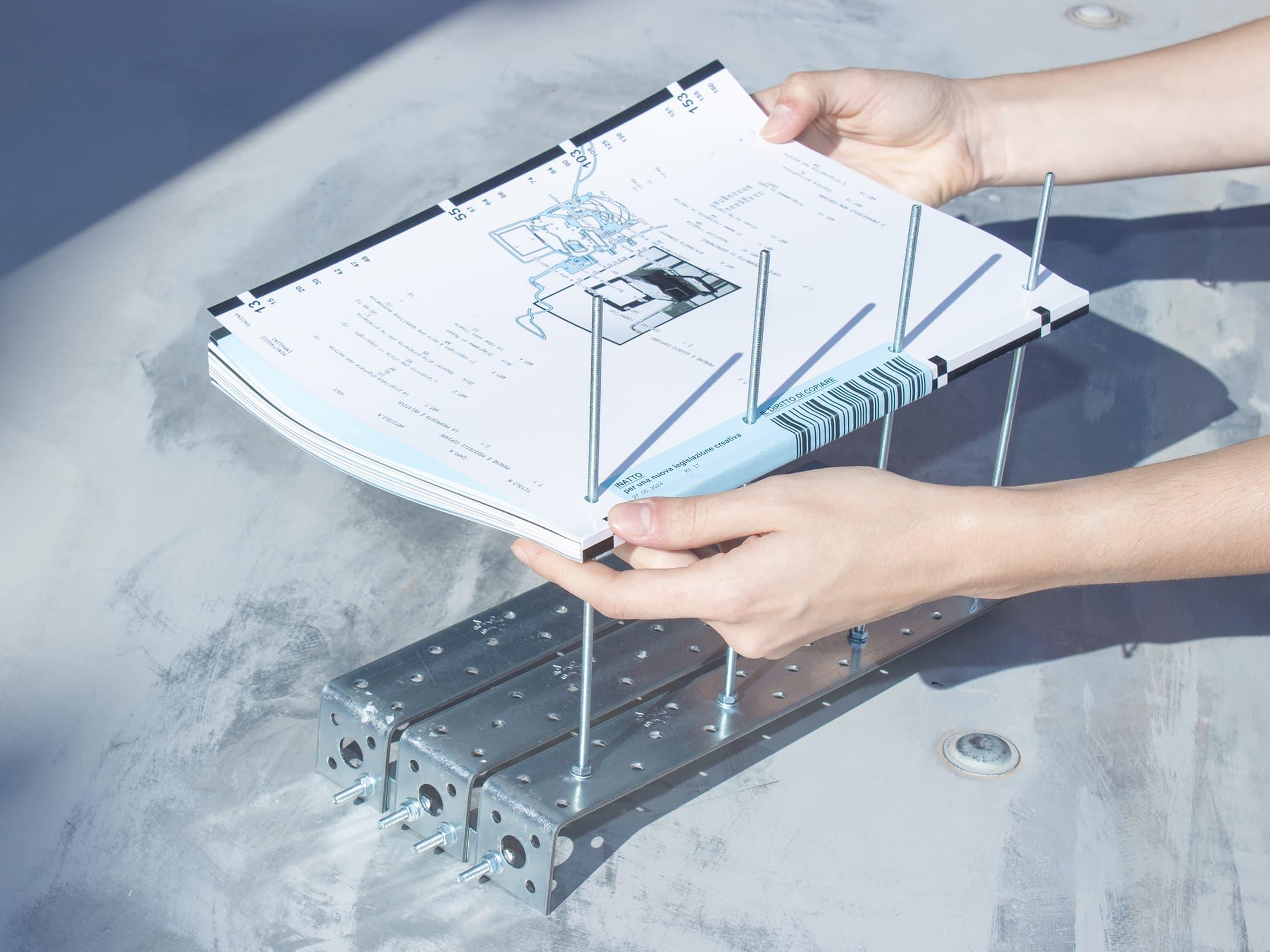 Hands holding a blueprint-like booklet with a small metal construction.