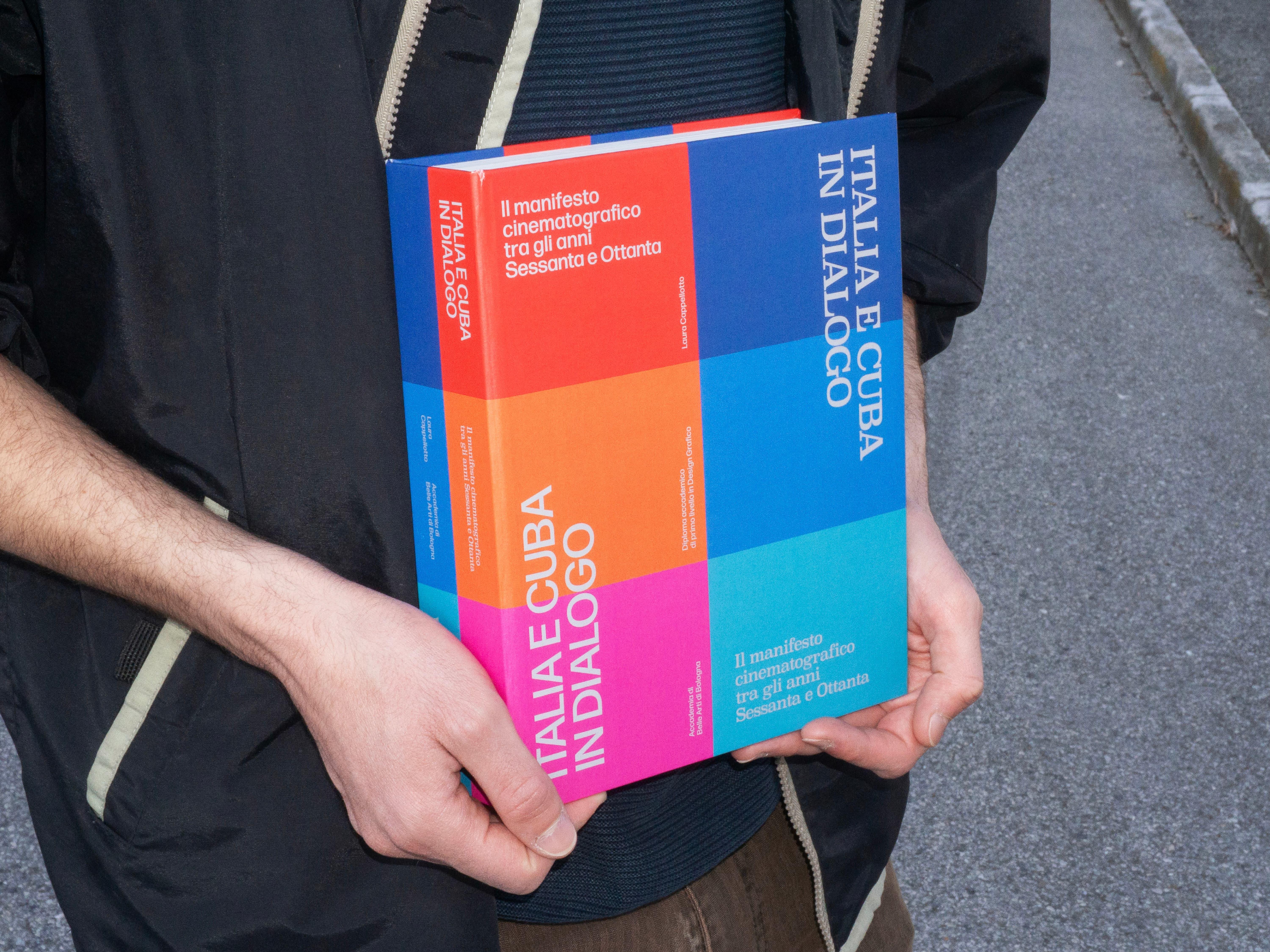 A person in a dark jacket holds the book Italia e Cuba in dialogo against their body. The cover and spine are made of bold blocks of red, blue, orange, magenta and turquoise, with the title in large white capitals.