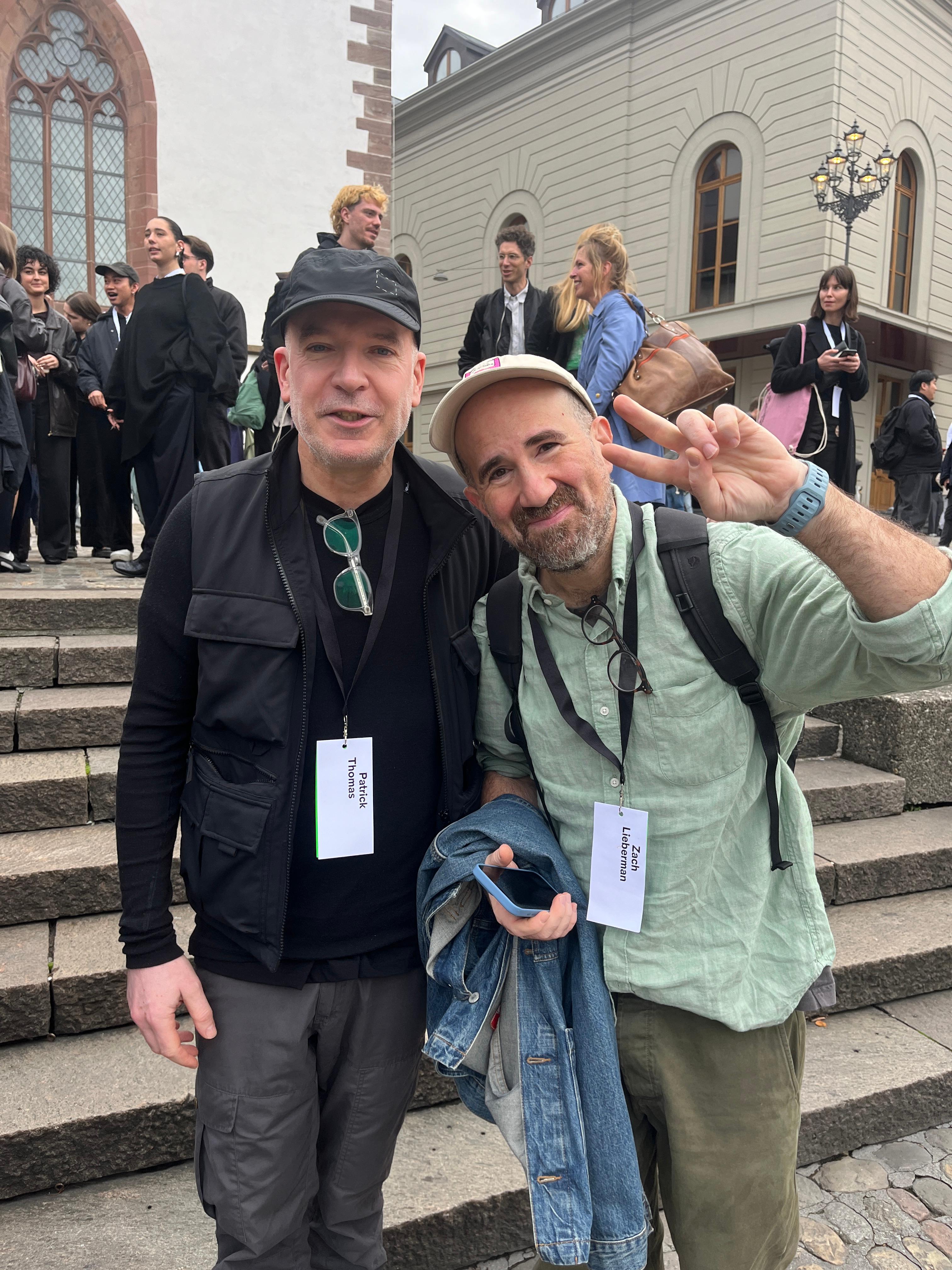 Two men wearing AGI Basel badges on steps outside a building.