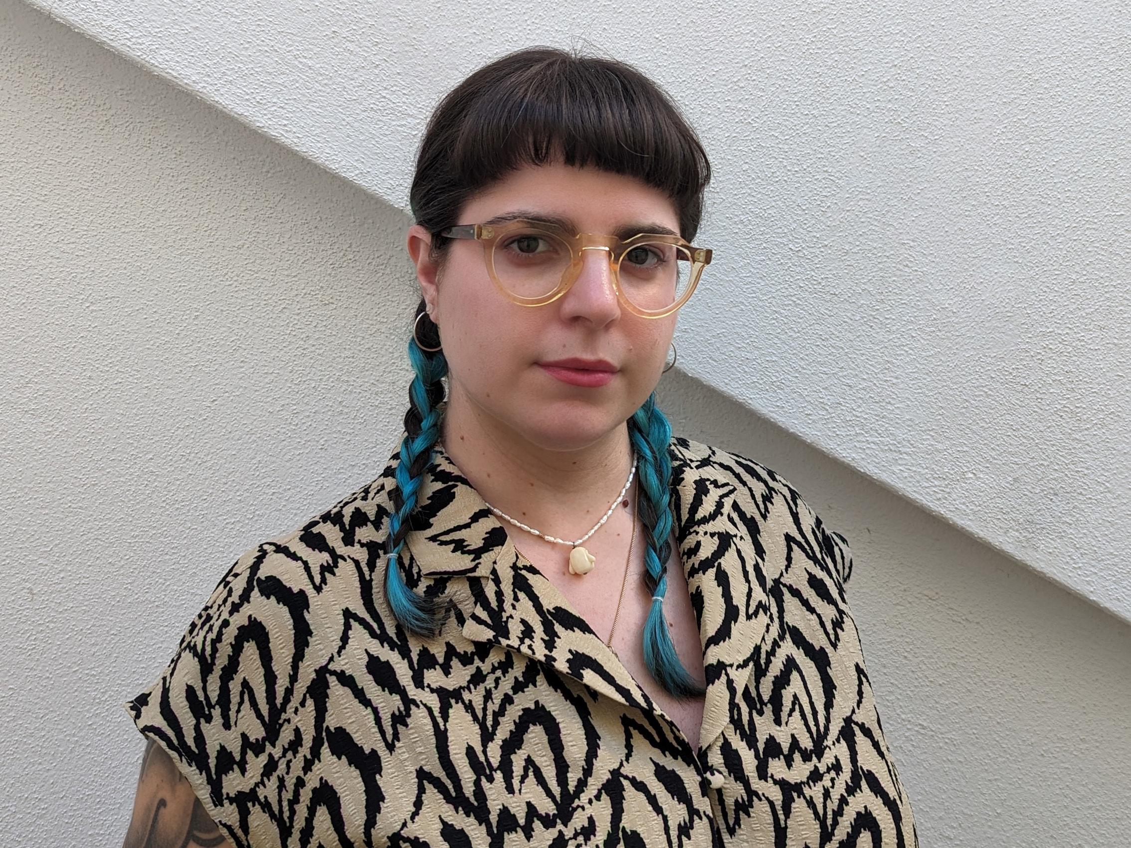 Portrait of a person with round yellow glasses and a patterned shirt against a pale wall.