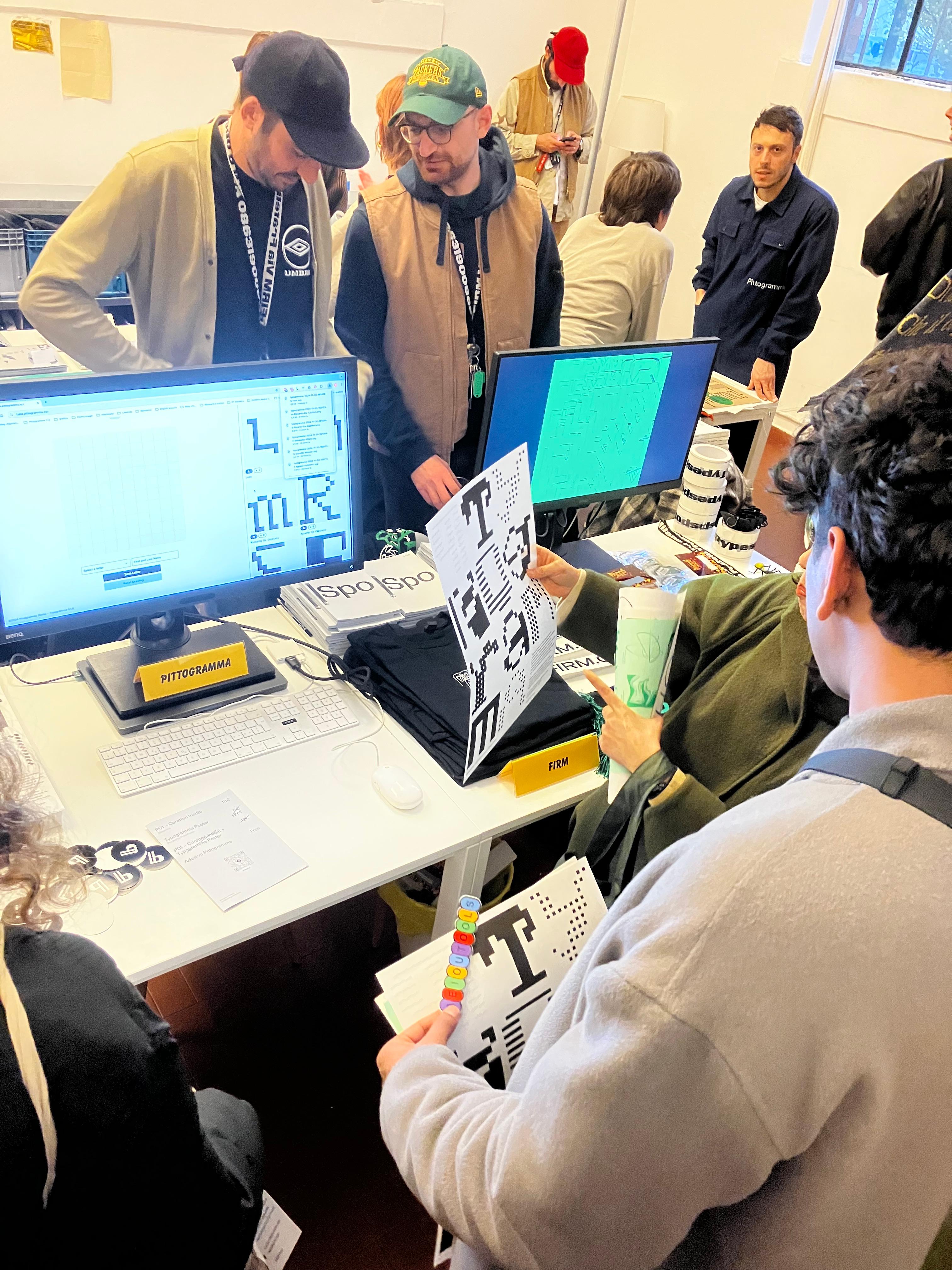 Attendees gathered around a table with computer screens, pixel-letter posters and printed matter.