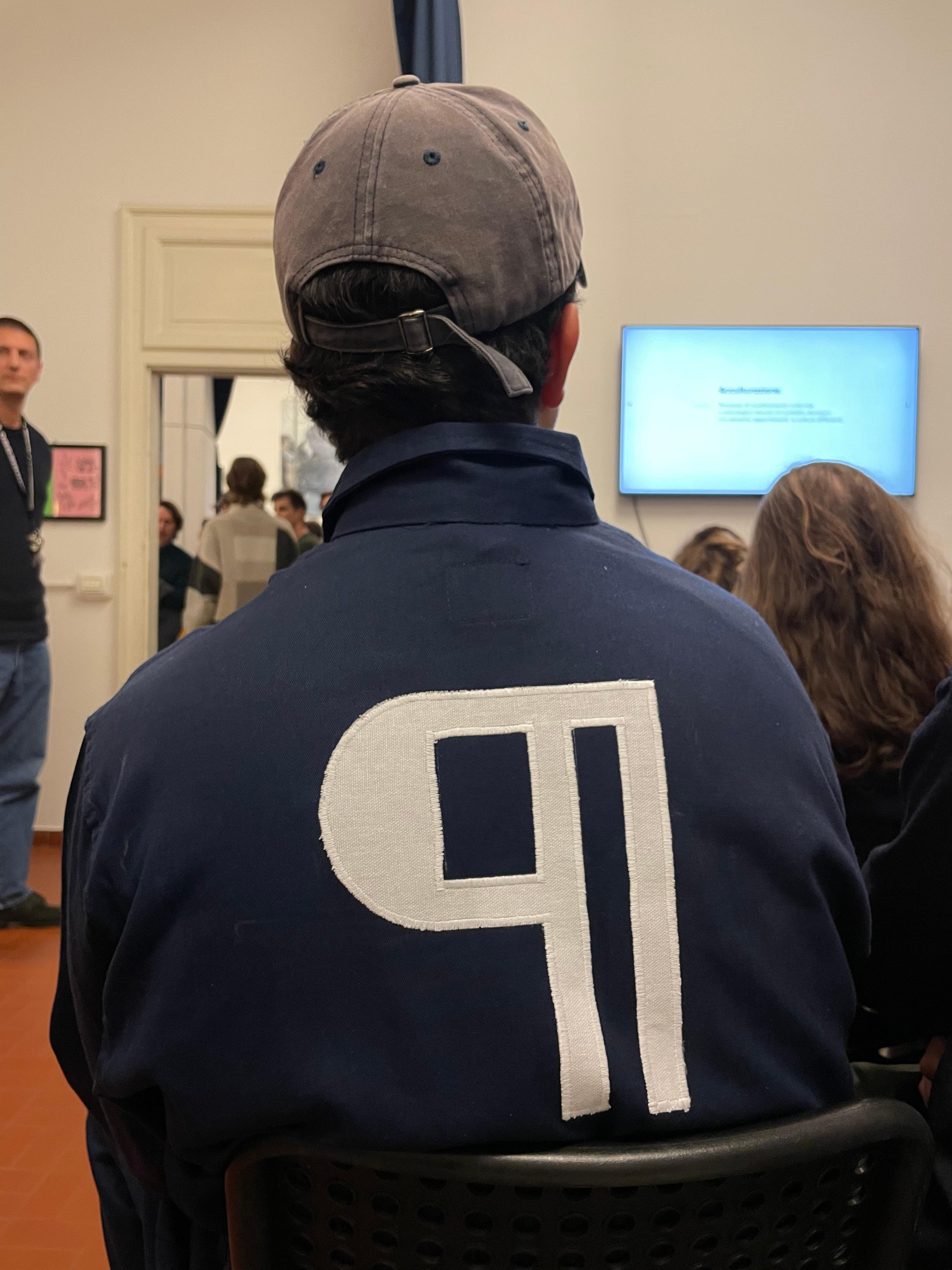 Back of an attendee wearing a jacket with a large white "91", facing a presentation screen.