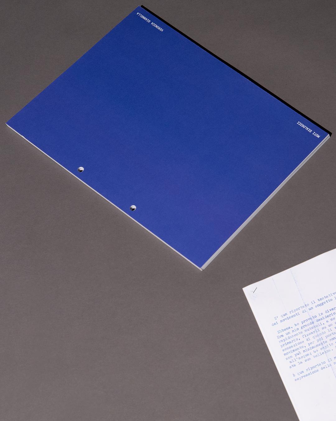 Closed blue Moti dialogici publication with a white text card on a gray surface.