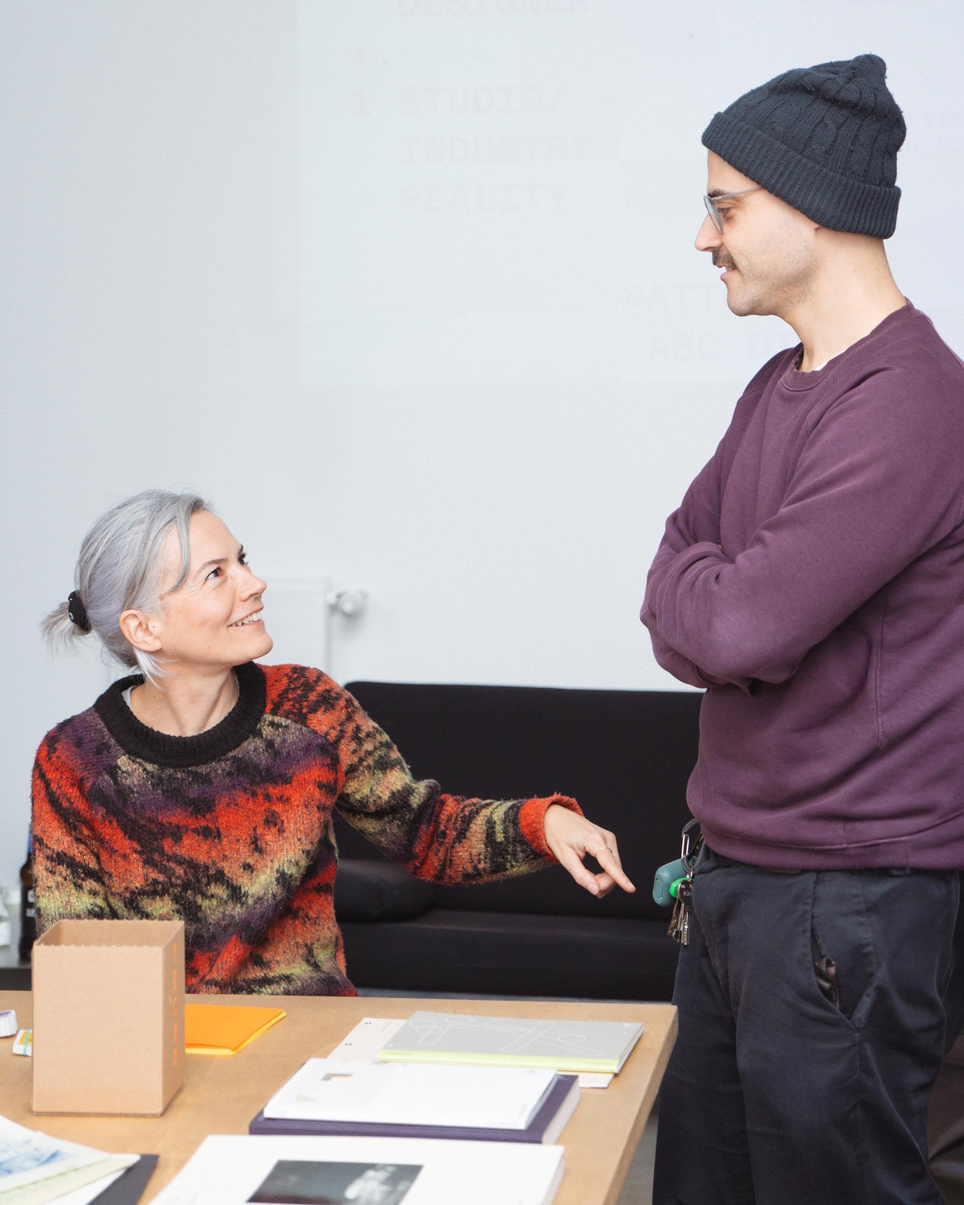 Two people talking across a table covered with books and papers.