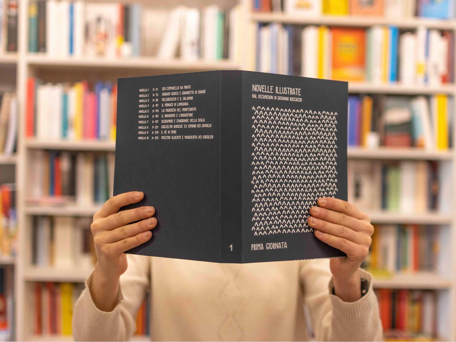 Person holding a black Novelle illustrate book in front of their face.