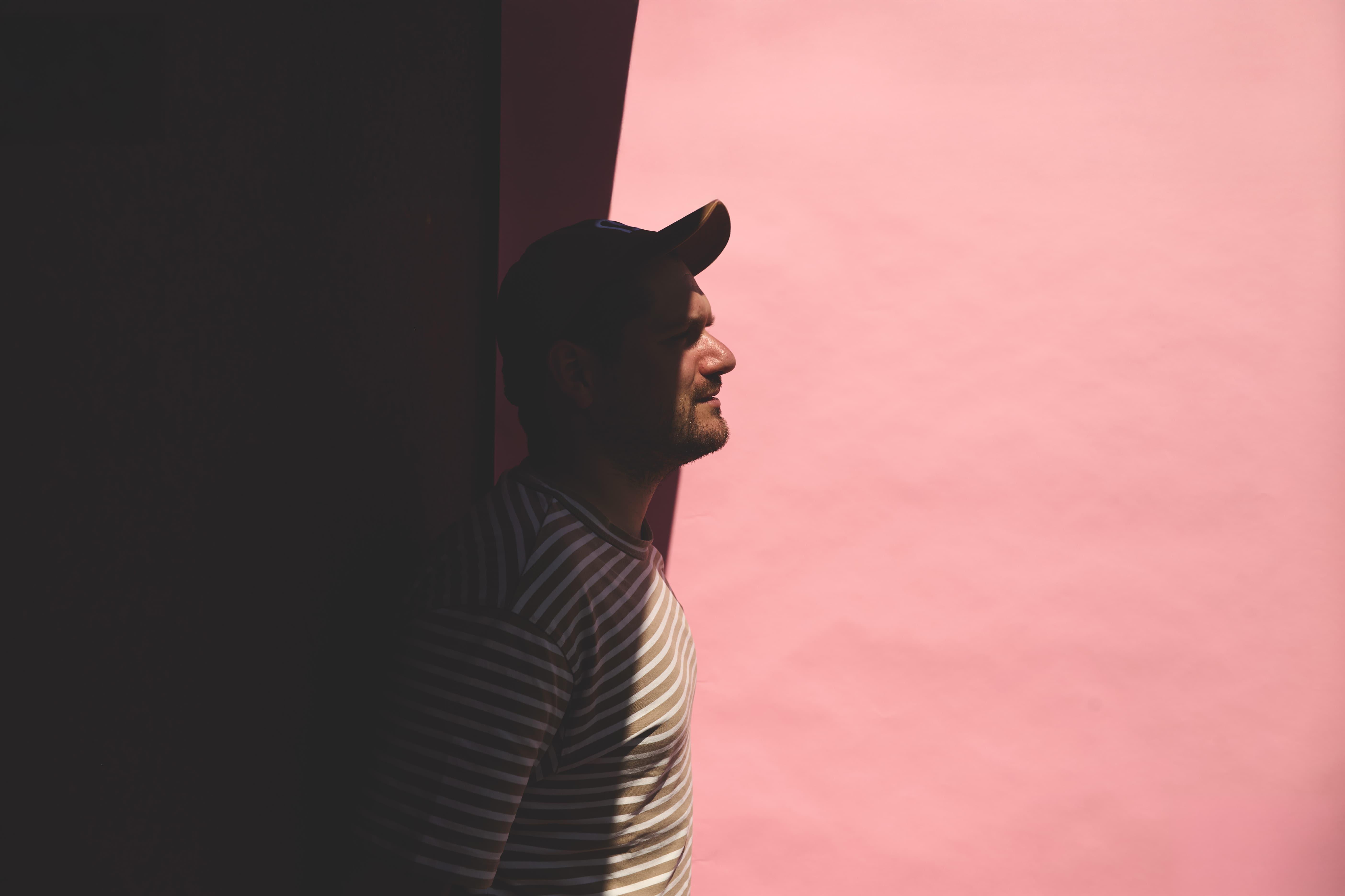 Person in a black cap and striped jacket standing beside a pink wall.
