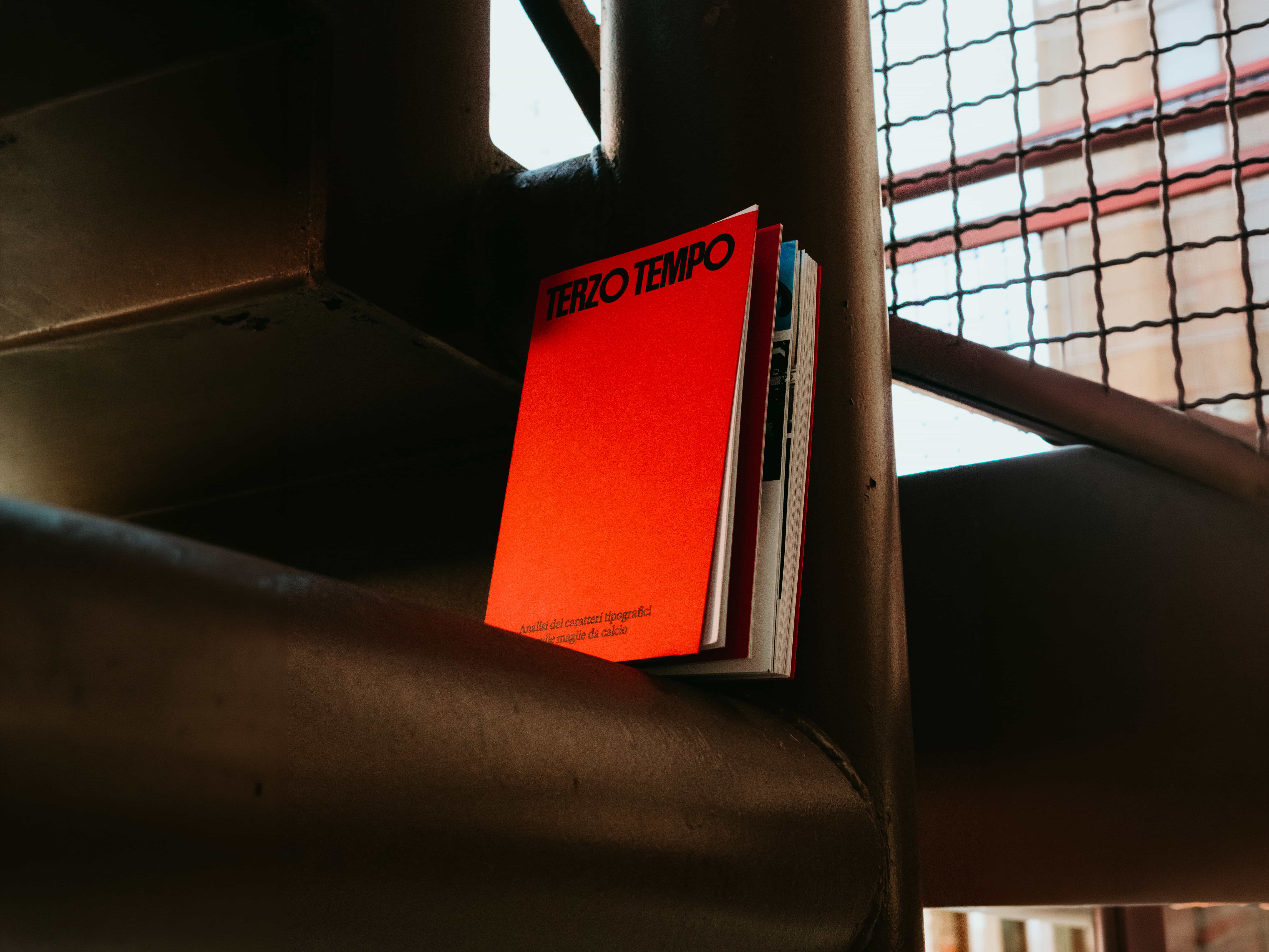 Terzo Tempo, a book with a bright red cover and bold black title, standing on a dark metal stair beam beside a mesh railing.