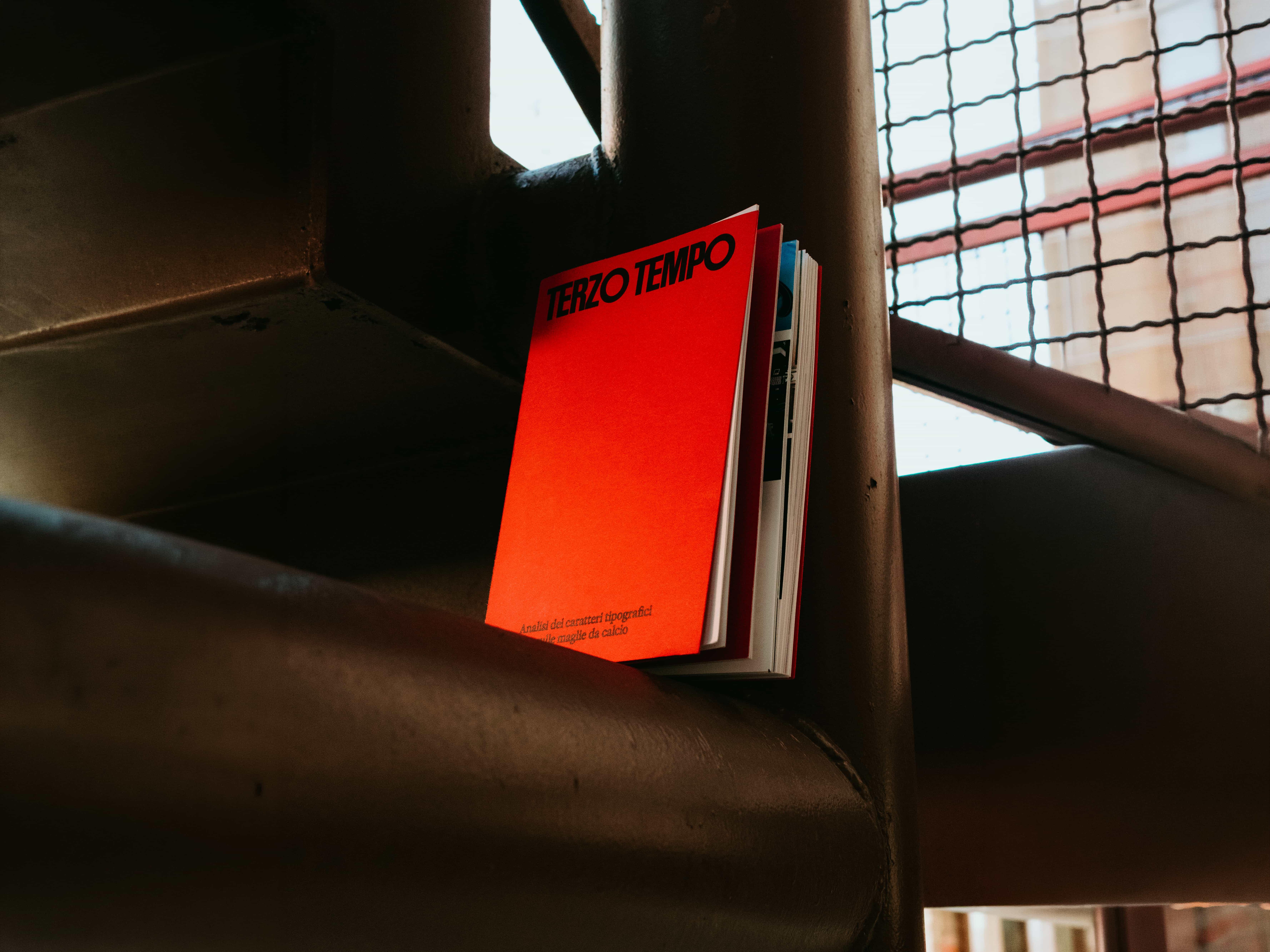 Terzo Tempo, a book with a bright red cover and bold black title, standing on a dark metal stair beam beside a mesh railing.