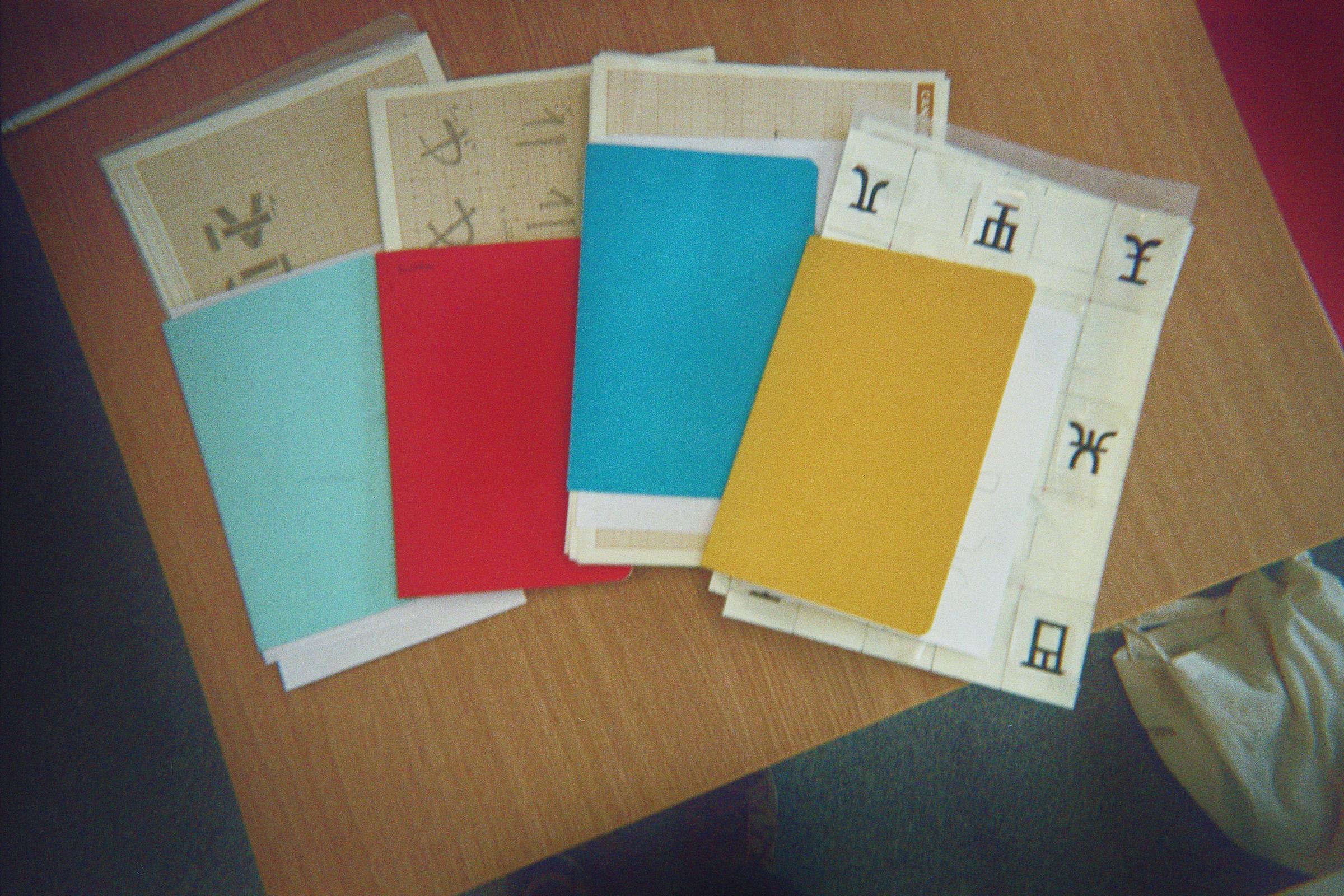 Coloured notebooks and paper sheets with glyph studies on a workshop table.