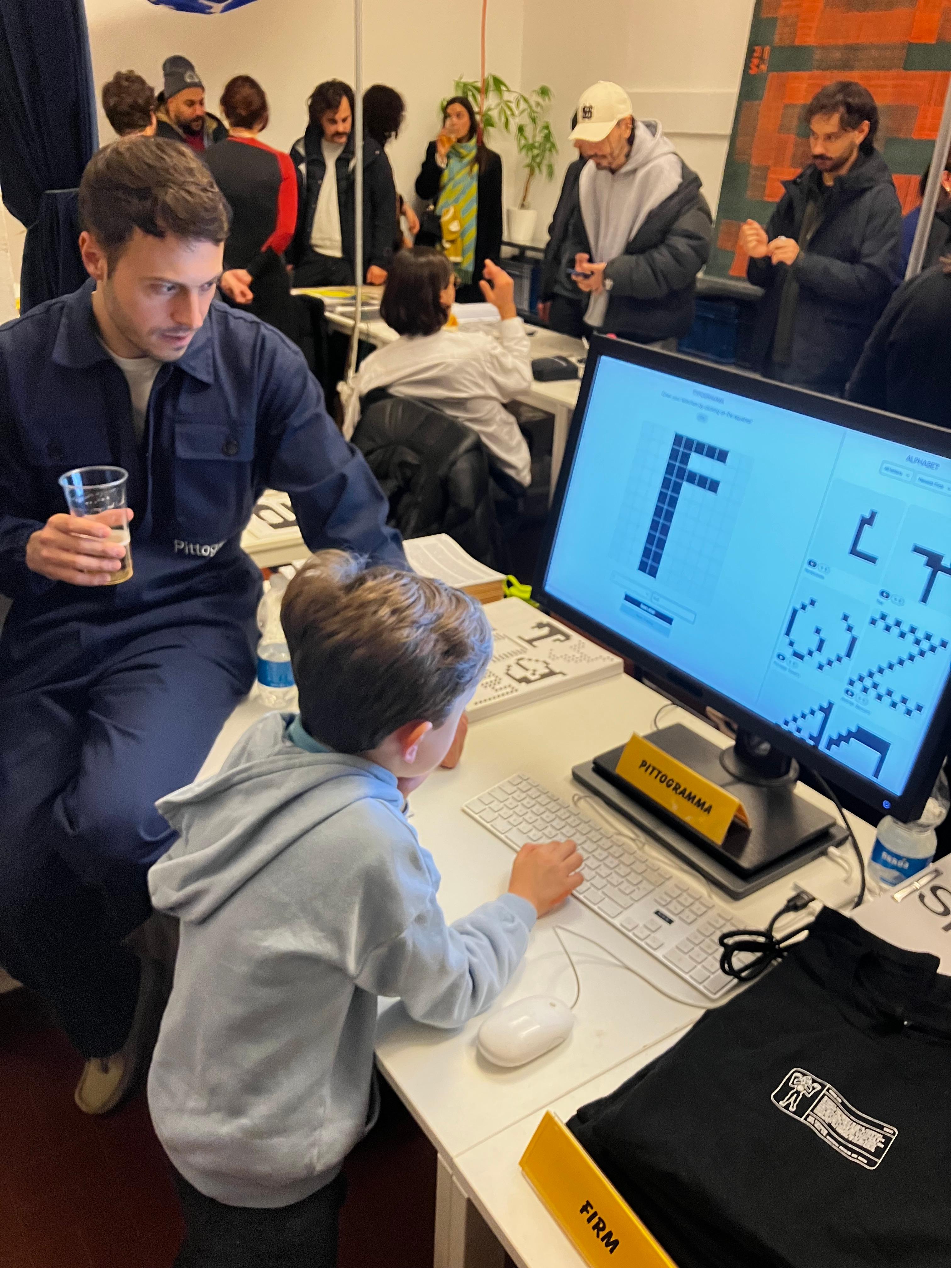 Child using a desktop computer with pixel-letter graphics at a crowded event table.