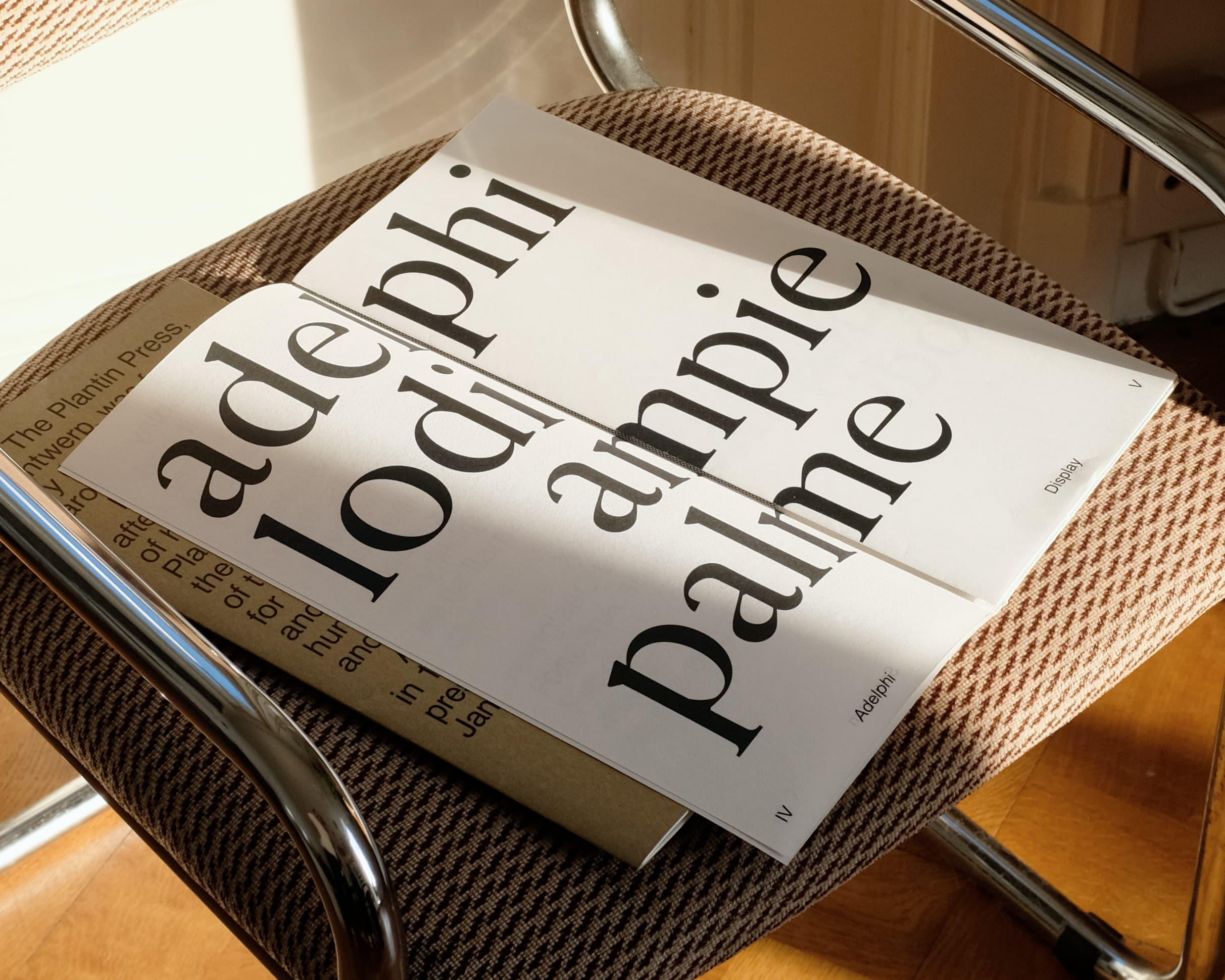 Open Adelphi publication with oversized black serif lettering on a patterned chair.