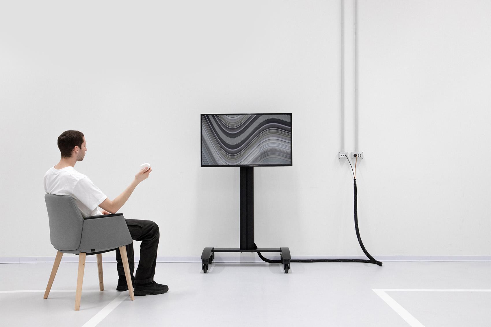 Person seated in a white room looking at a black screen on a stand.