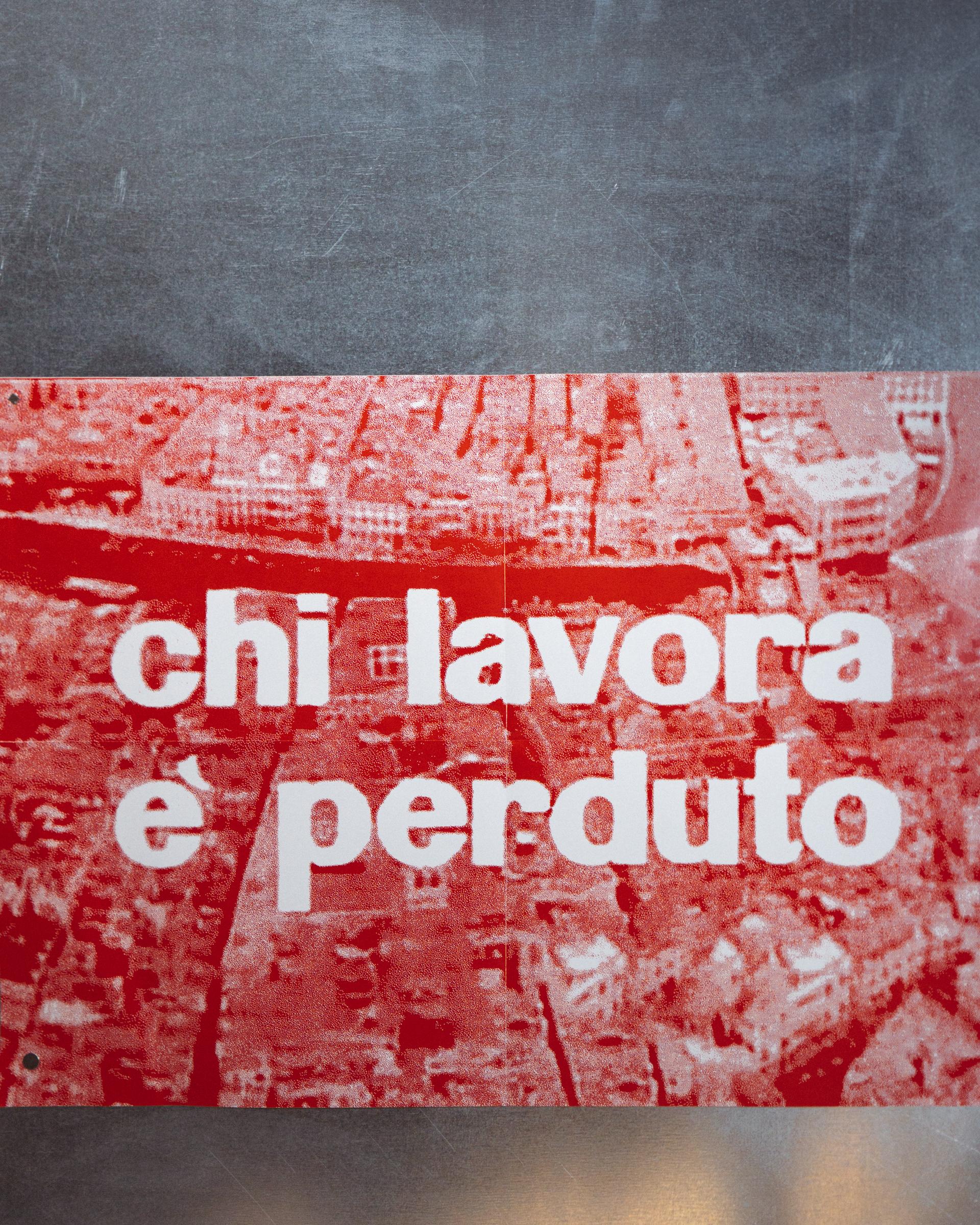 Red poster reading "chi lavora e perduto" on a grey wall.