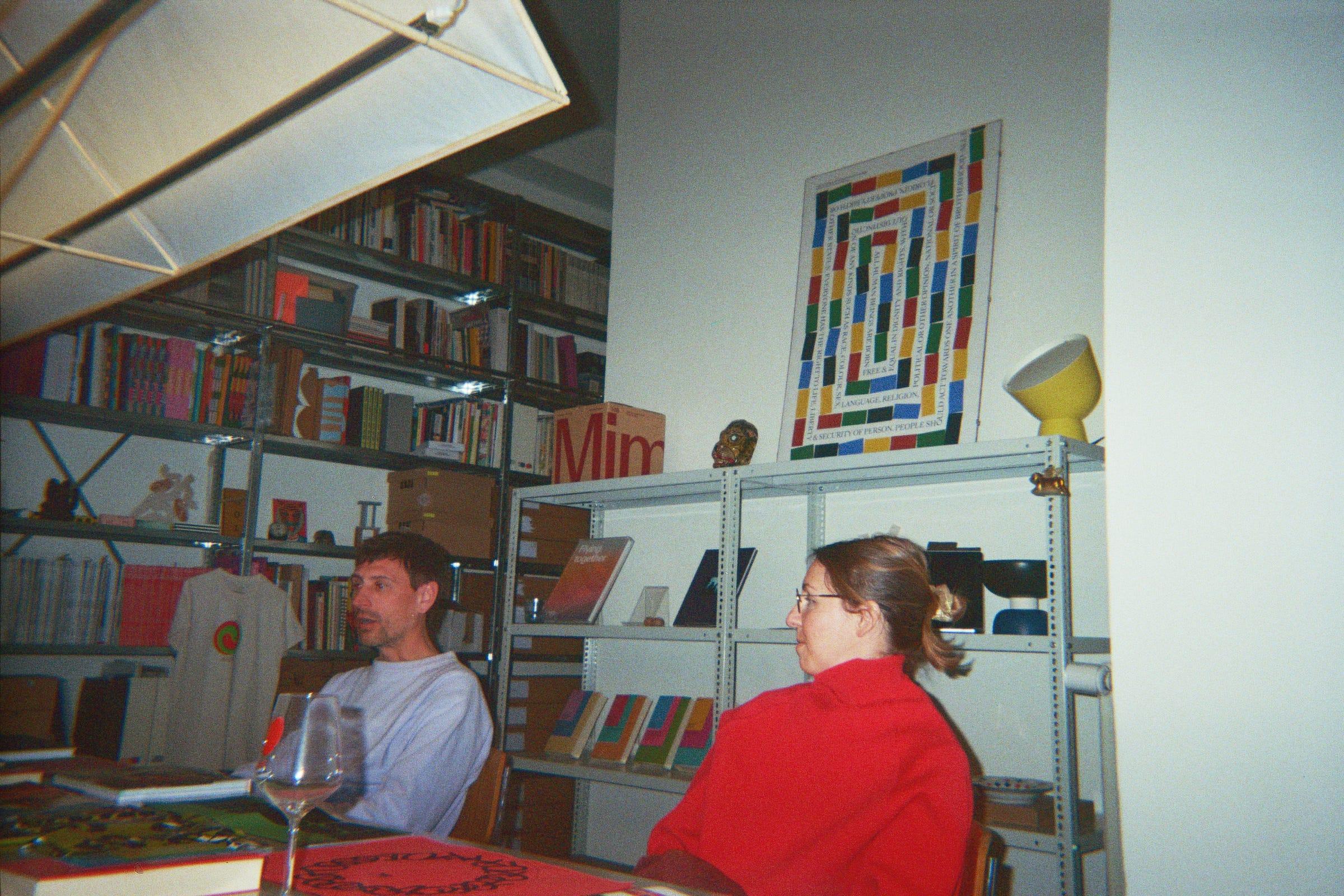 Two people seated in front of LaTigre's shelves of books, objects and printed work.