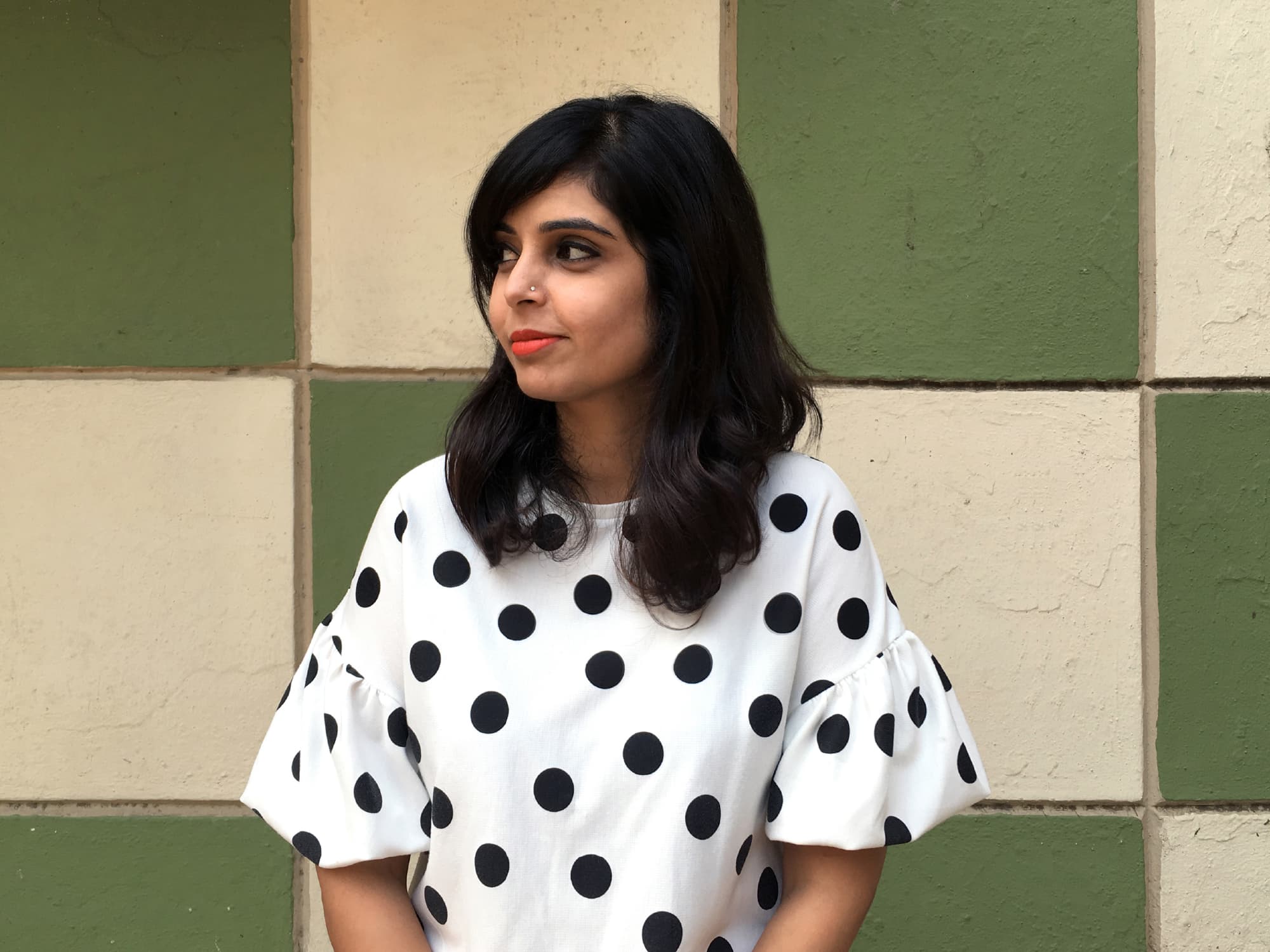 Portrait of a person in a white shirt with black polka dots against a green tiled wall.