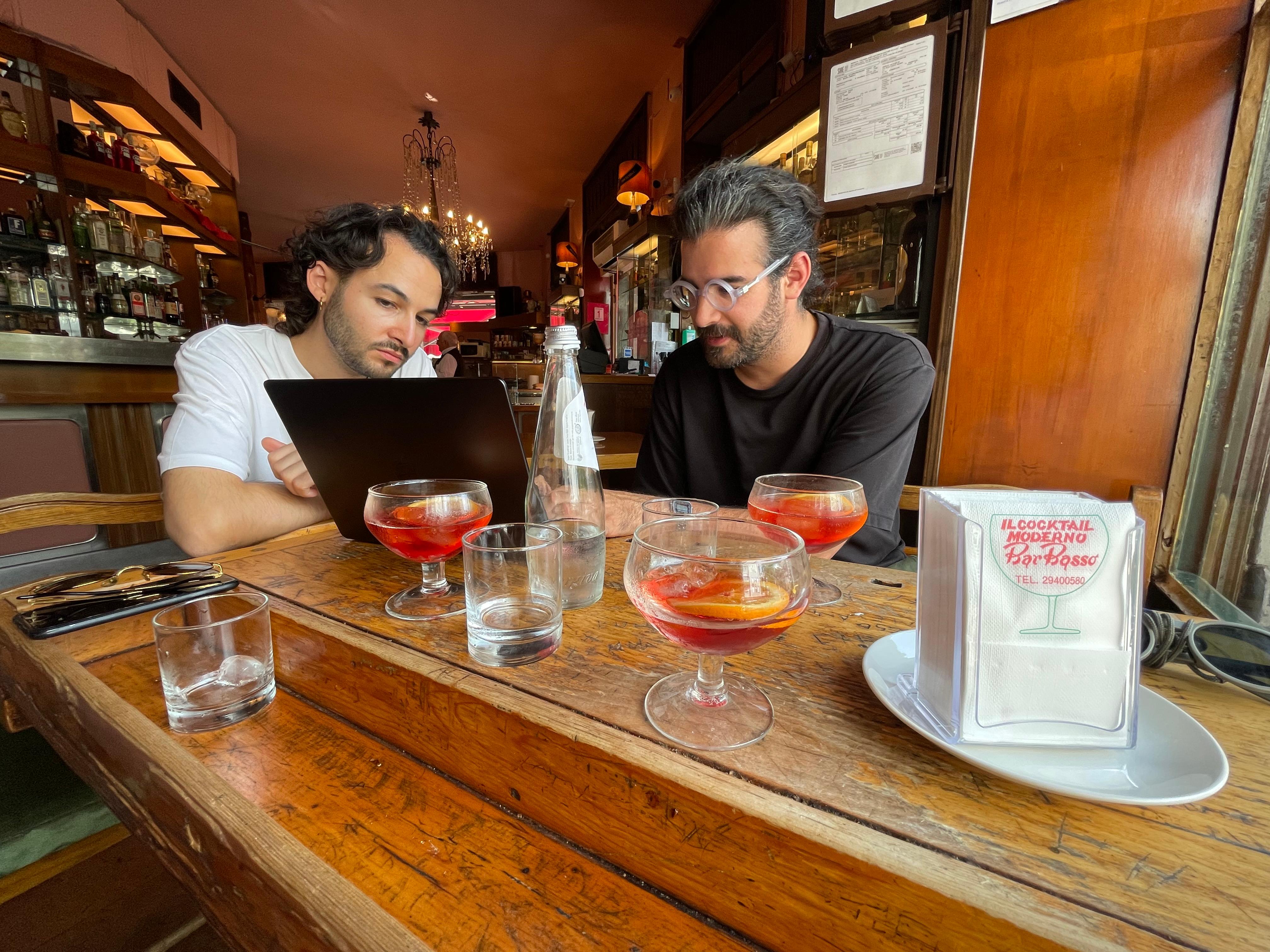 Two people working on a laptop at a cafe table with aperitifs.