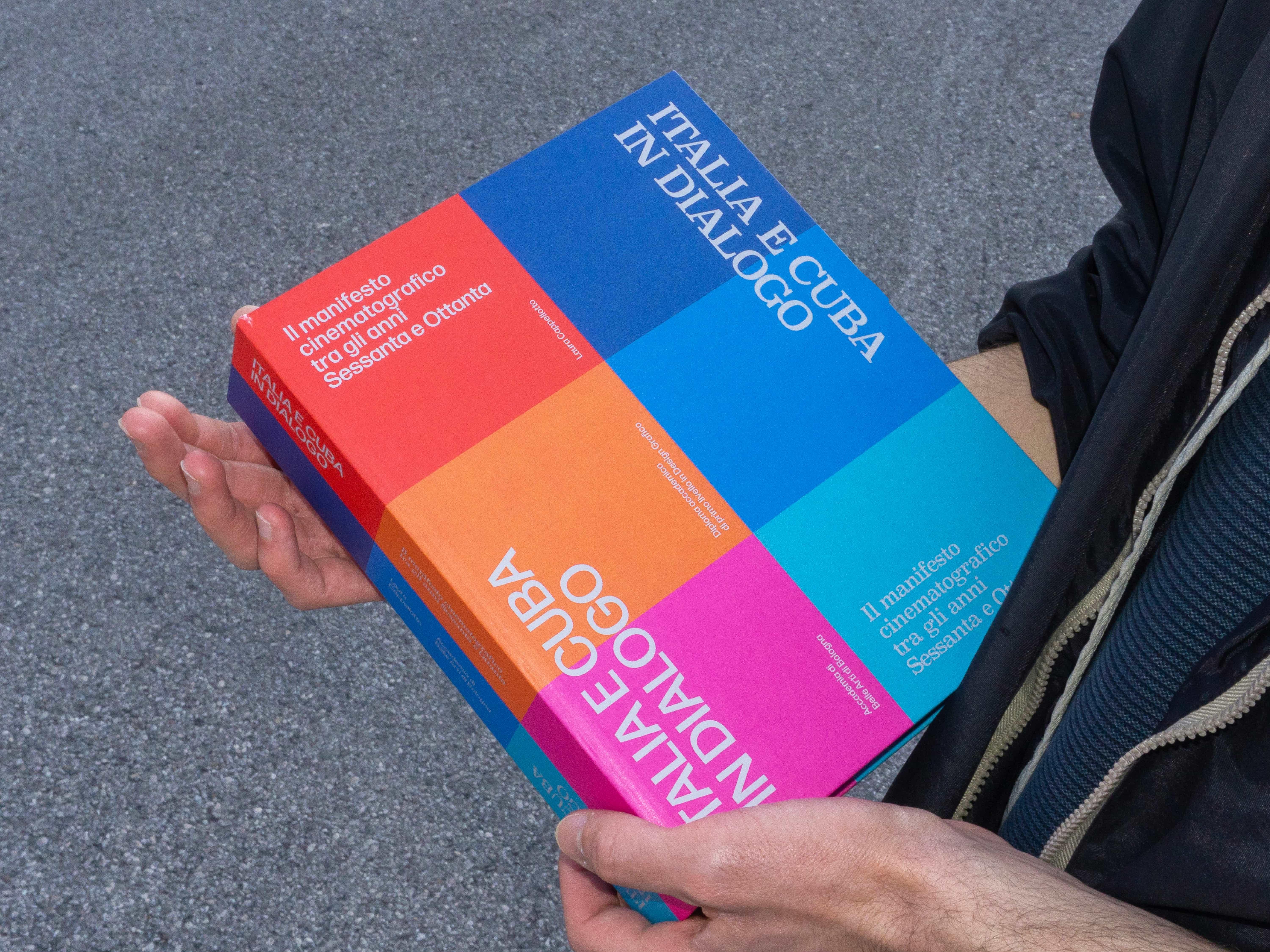 Hands holding Italia e Cuba in dialogo, its cover made of red, blue, orange, magenta and turquoise blocks.
