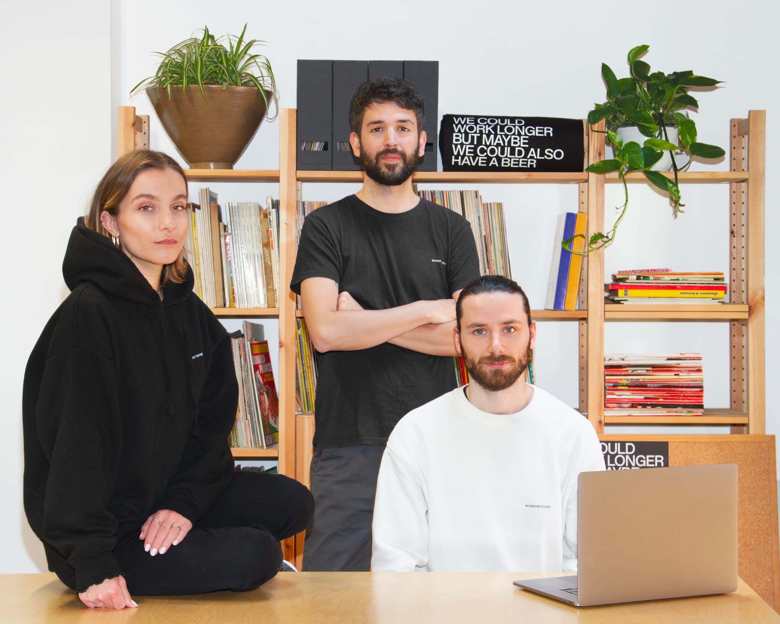 Three people in black and white clothing around a desk with a laptop, books and plants.