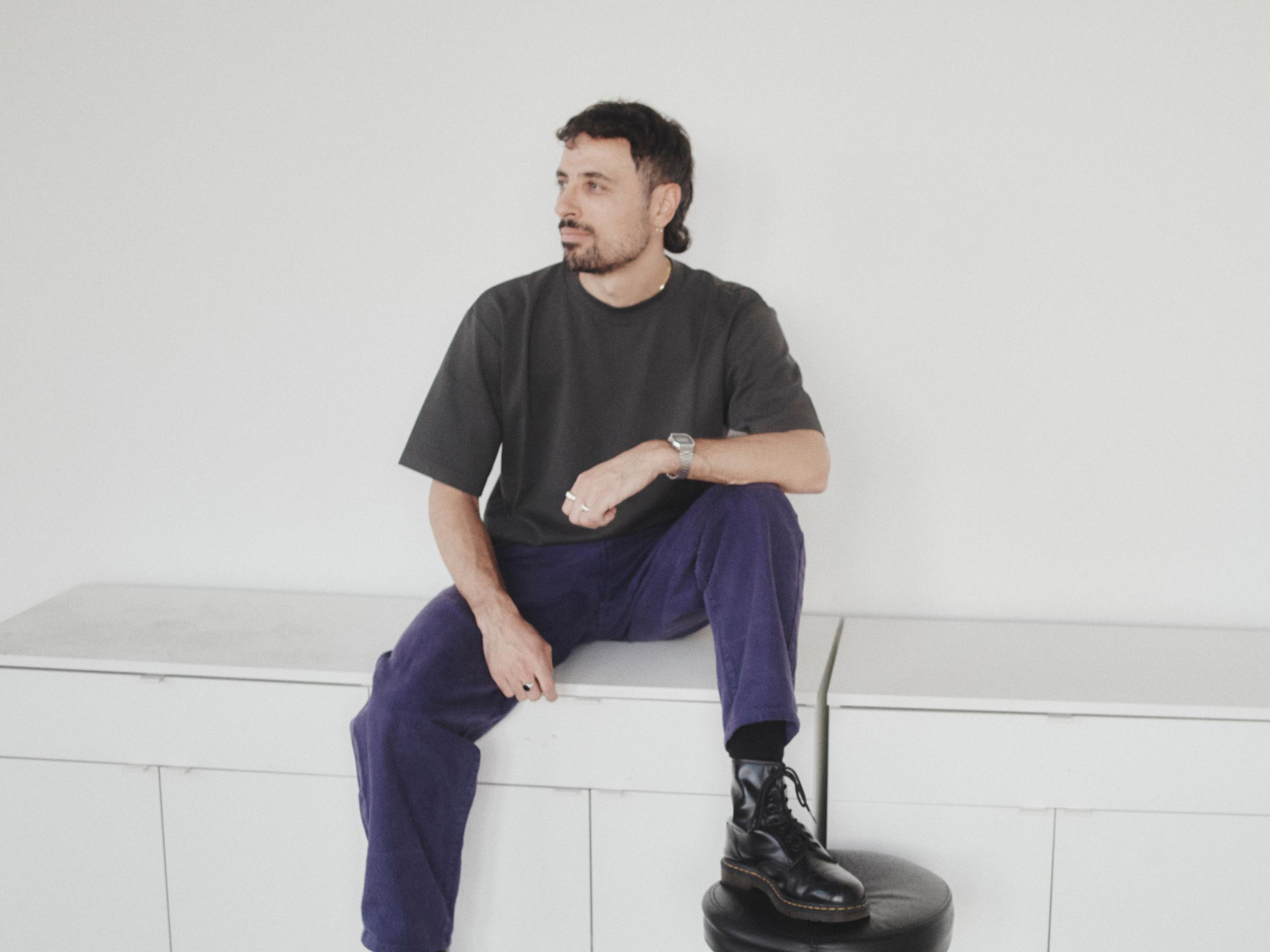 Vittorio Perotti sitting on a white cabinet in a grey T-shirt and purple trousers, looking off to the side.