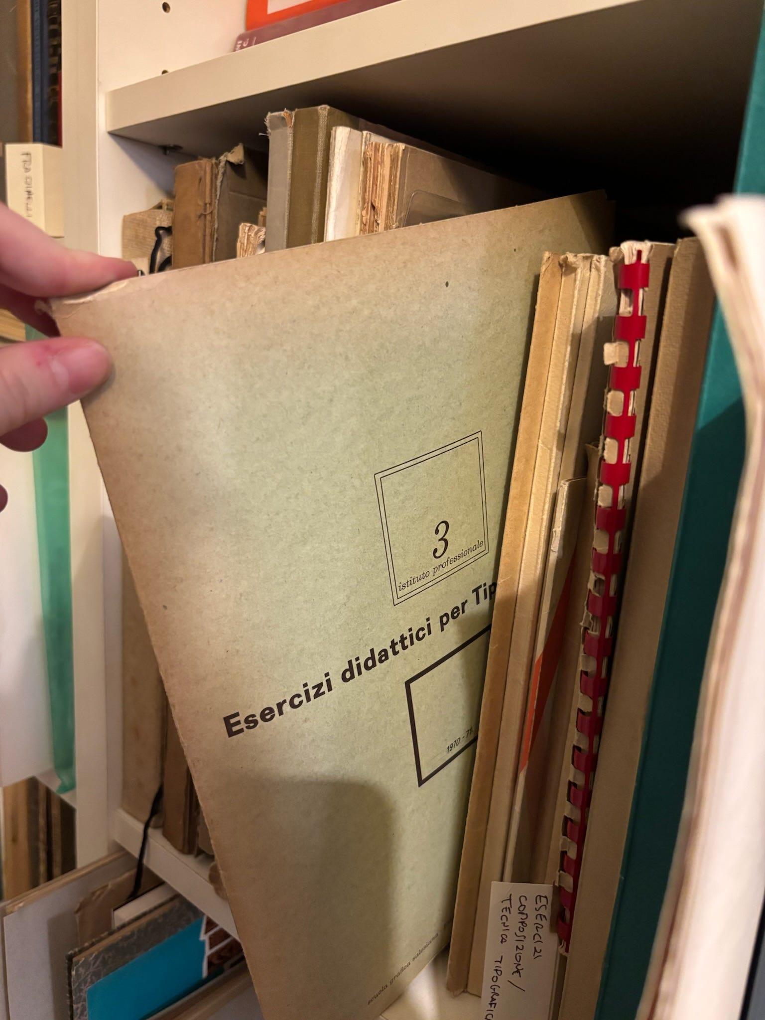 Hand pulling a worn "Esercizi didattici" exercise booklet from an archive shelf.