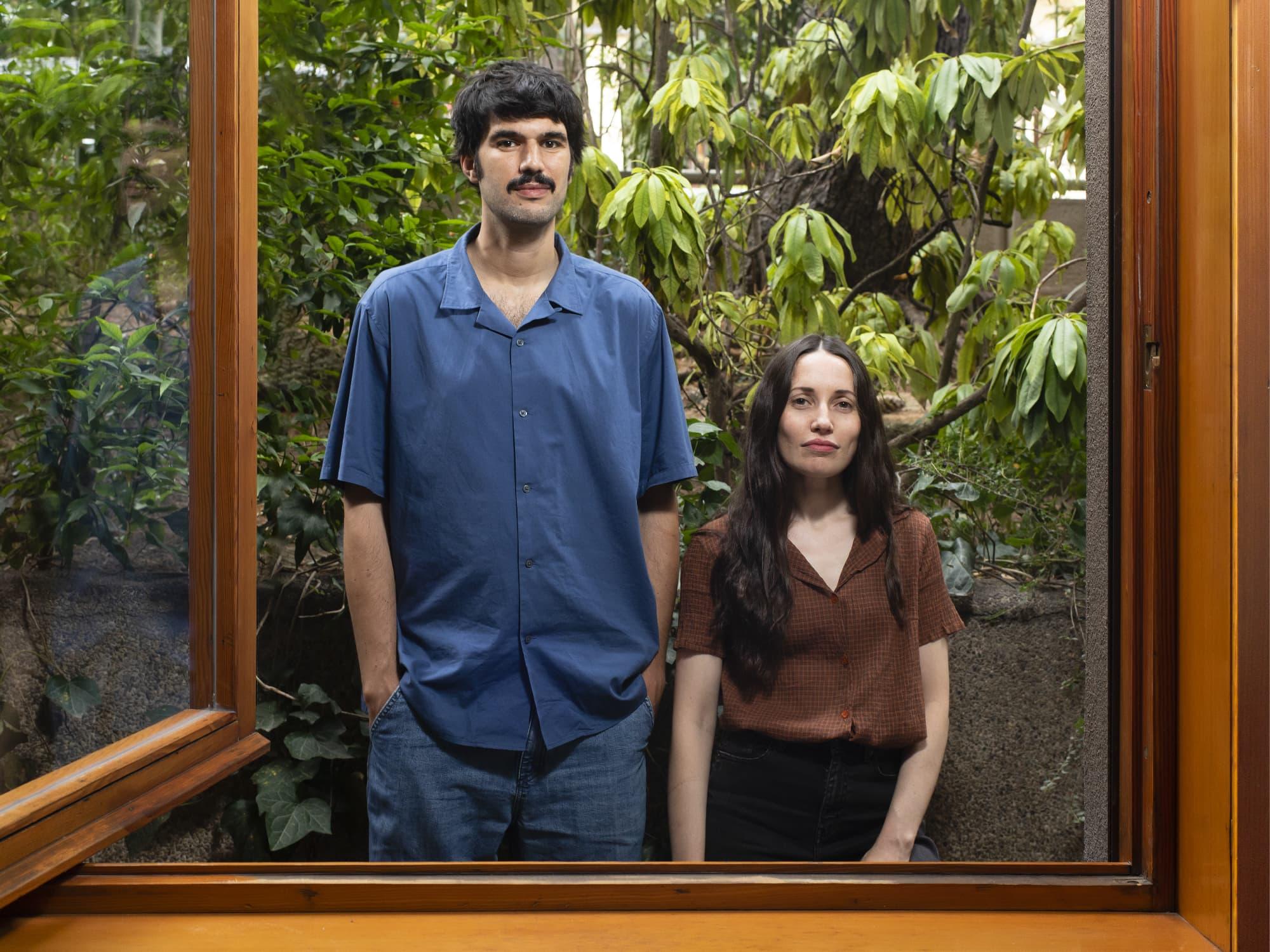 Two people standing in a wooden doorway with plants behind them.