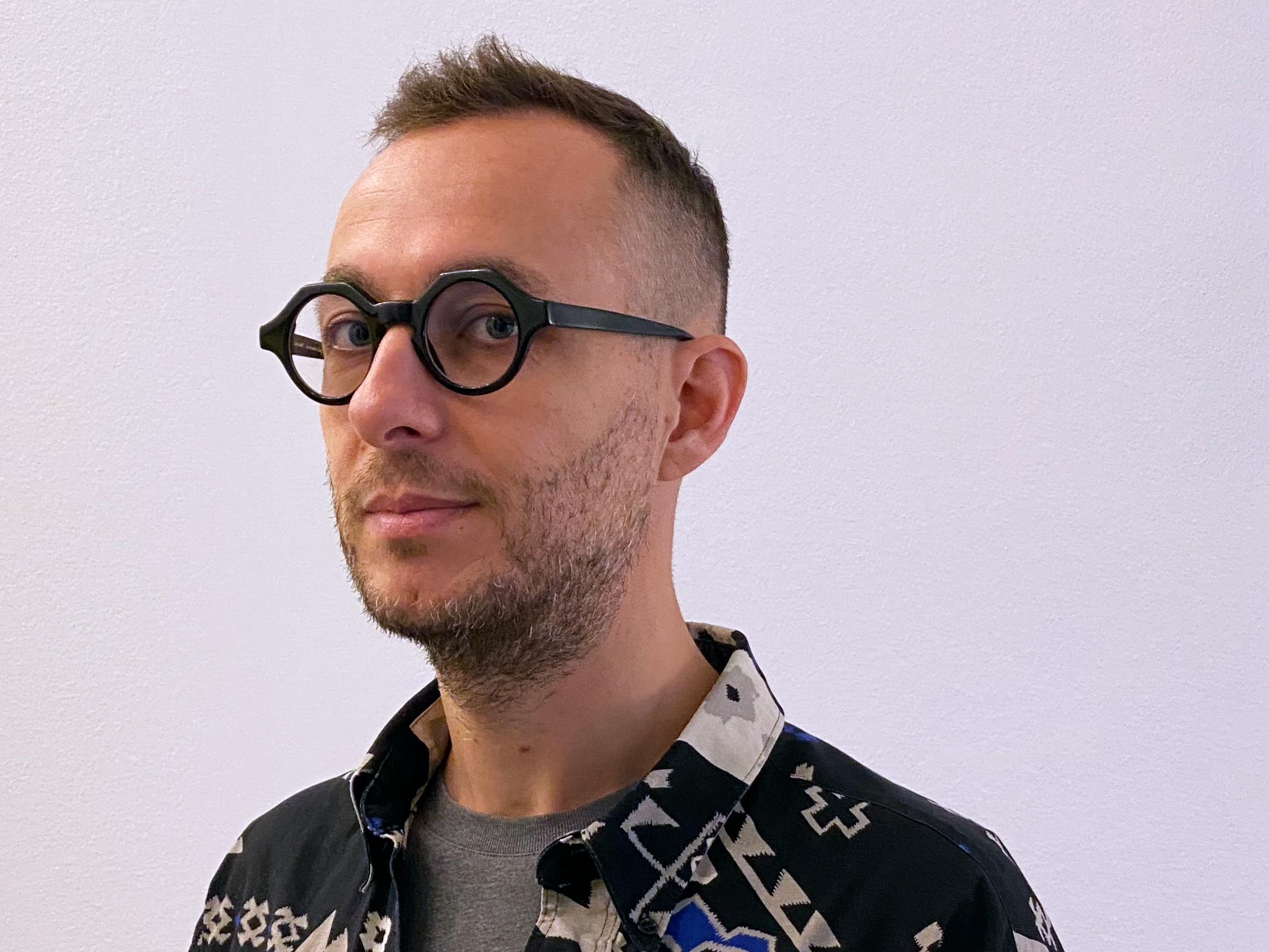 Portrait of a person wearing round black glasses and a patterned jacket against a pale background.