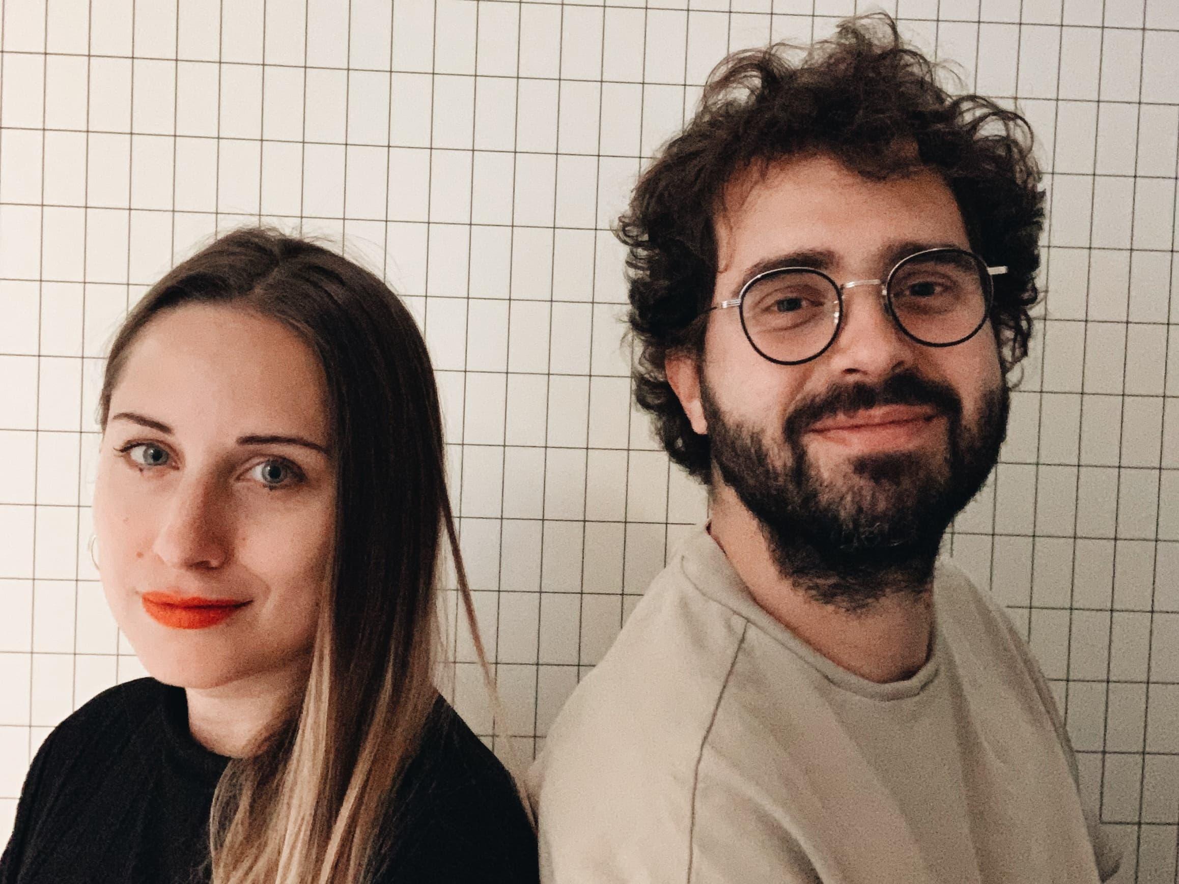 Two people standing side by side in front of a white tiled wall.