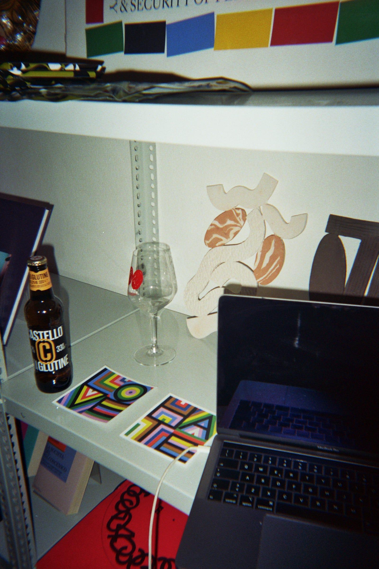 Laptop, beer bottle, printed card and a small animal cutout on a studio shelf.
