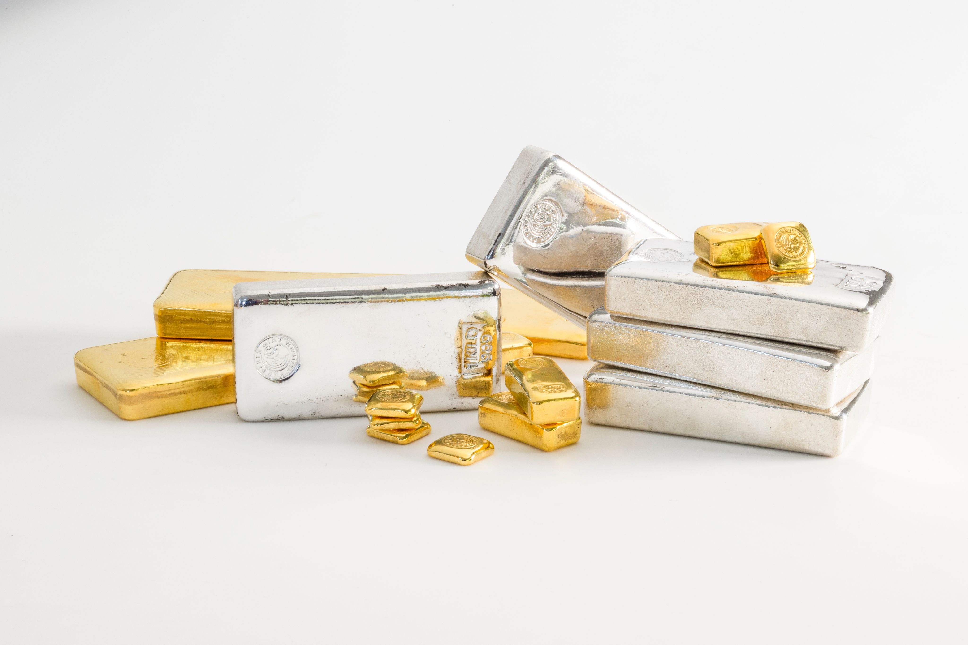 Stacks of shiny silver and gold bars arranged on a clean, white background. Smaller gold pieces are scattered in the foreground, suggesting wealth.
