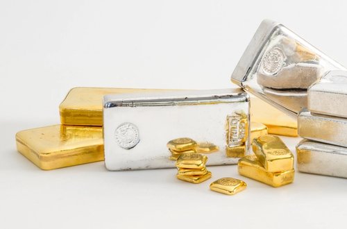 Stacks of shiny silver and gold bars arranged on a clean, white background. Smaller gold pieces are scattered in the foreground, suggesting wealth.
