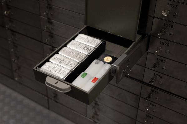 Open safety deposit box displaying five silver bars, two labeled tubes, and a single gold coin. The background shows a row of secure, locked boxes.