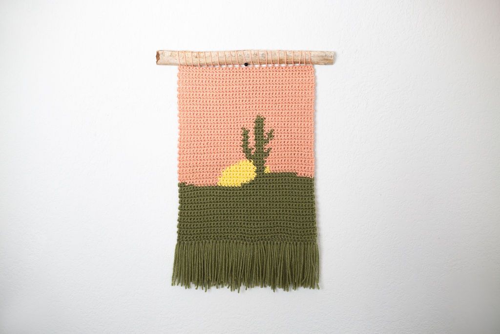 Crochet tapestry of a saguaro cactus against a peach sky over green ground