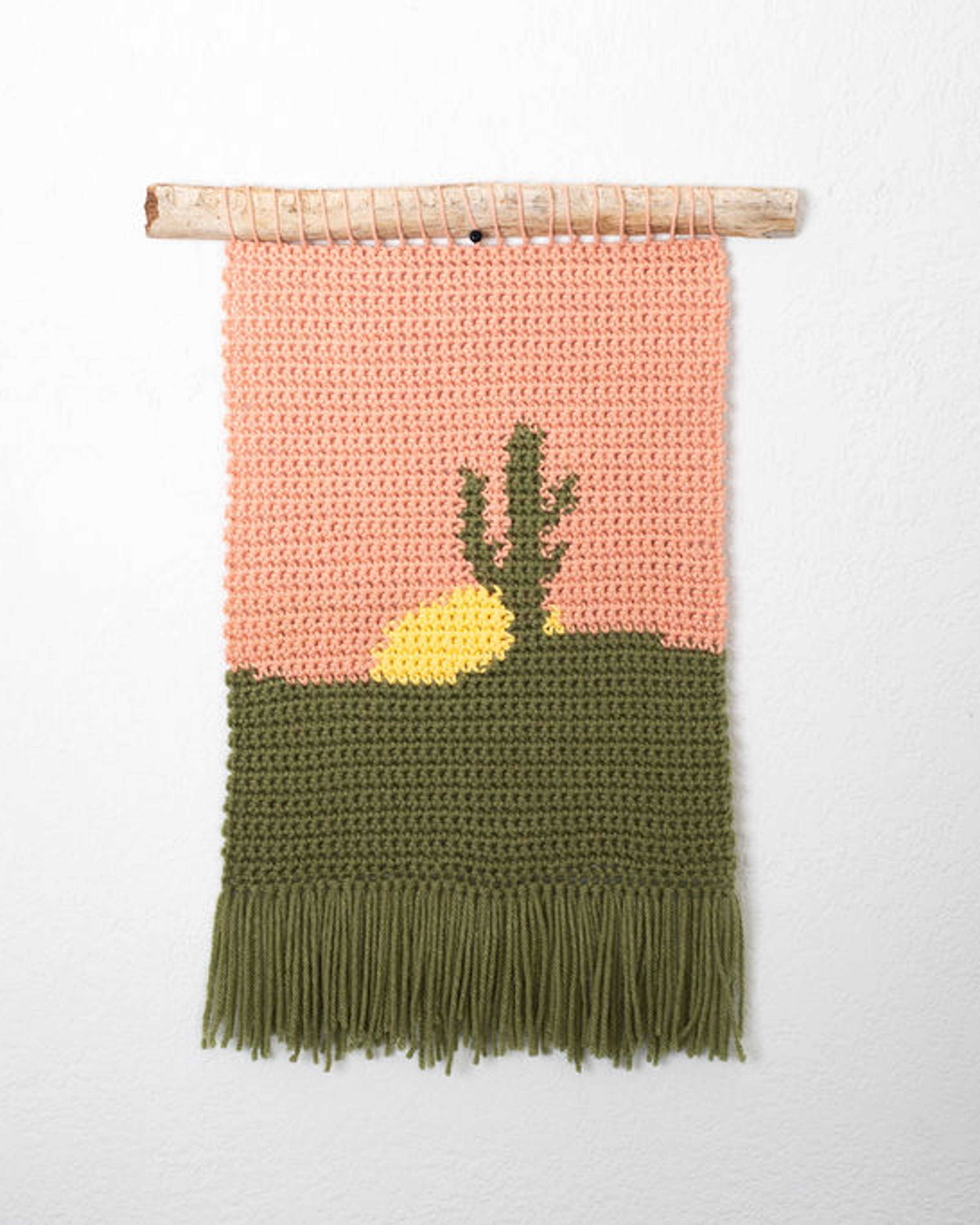 Crochet tapestry of a saguaro cactus against a peach sky over green ground