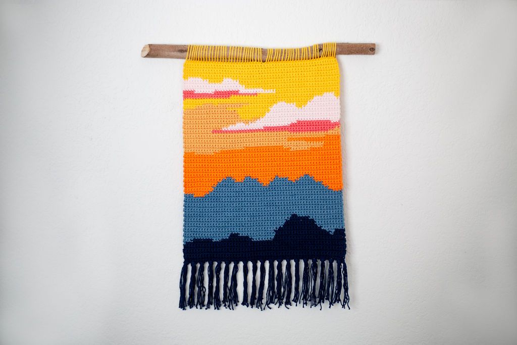 Crochet tapestry of a desert sunset in yellow, orange and pink over dark blue mountains, with navy fringe