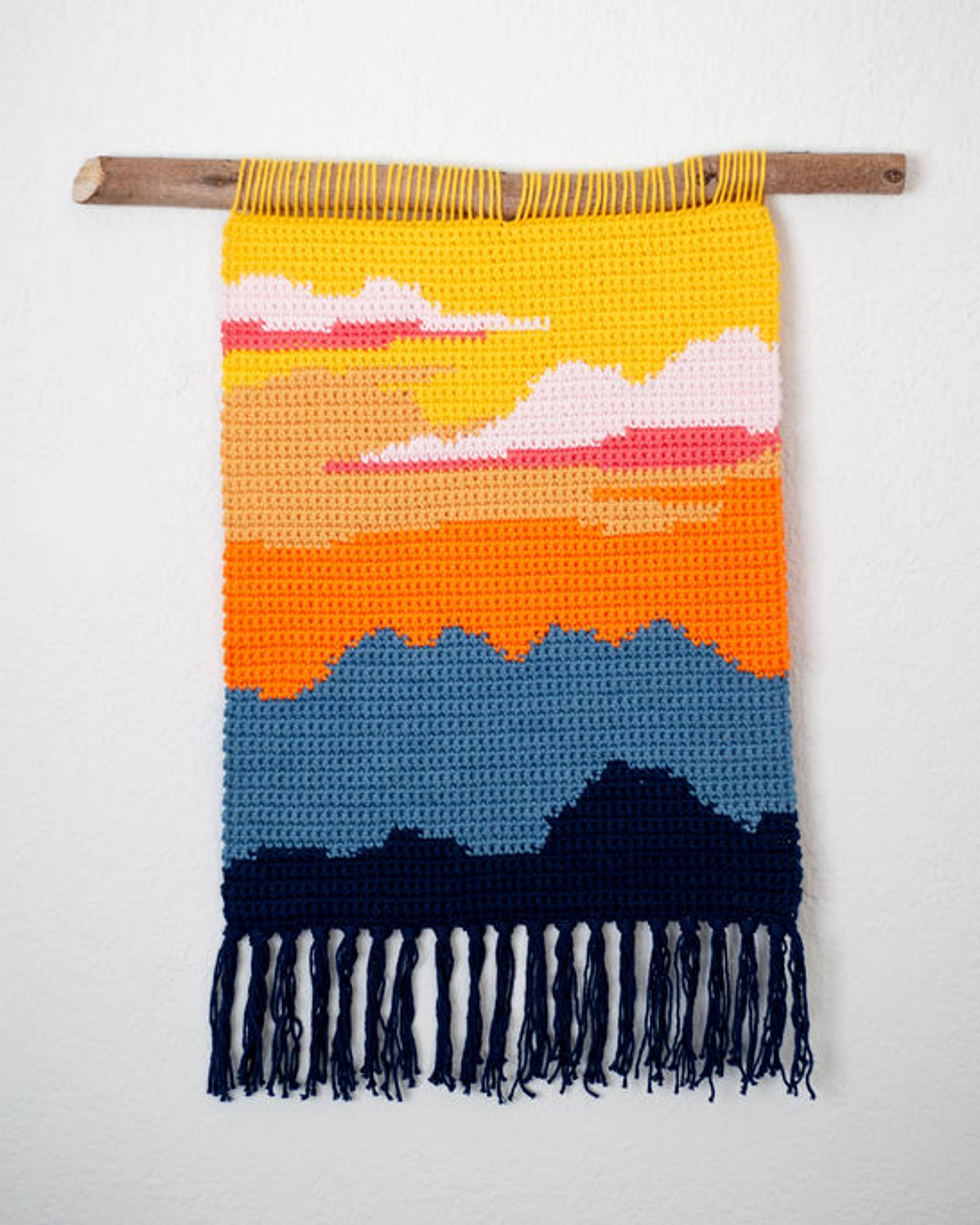 Crochet tapestry of a desert sunset in yellow, orange and pink over dark blue mountains, with navy fringe