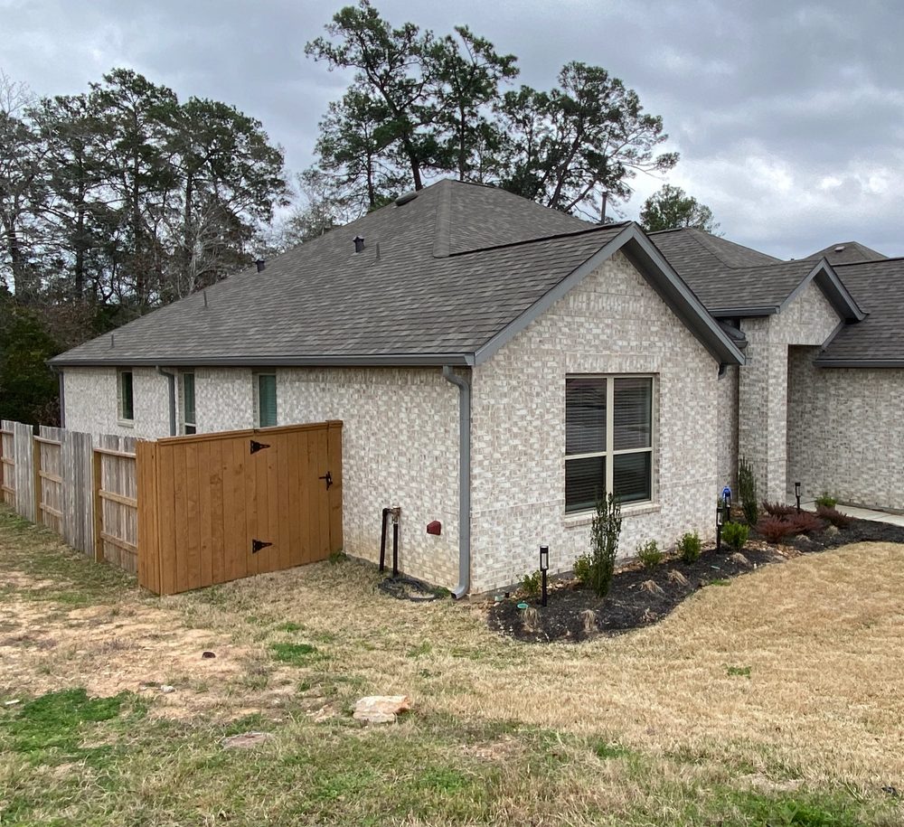 Dark gray seamless gutters and downspouts wrapping the light-brick front and side of a Conroe home