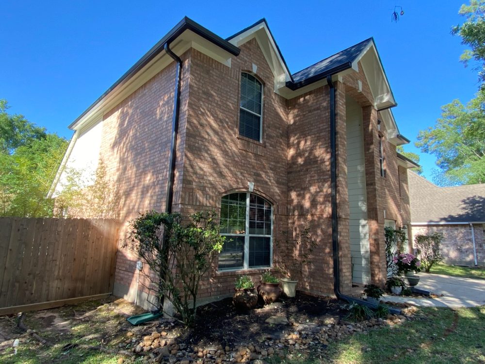 Dark bronze gutters and tall matching downspouts on the front corner of a two-story brick home in The Woodlands