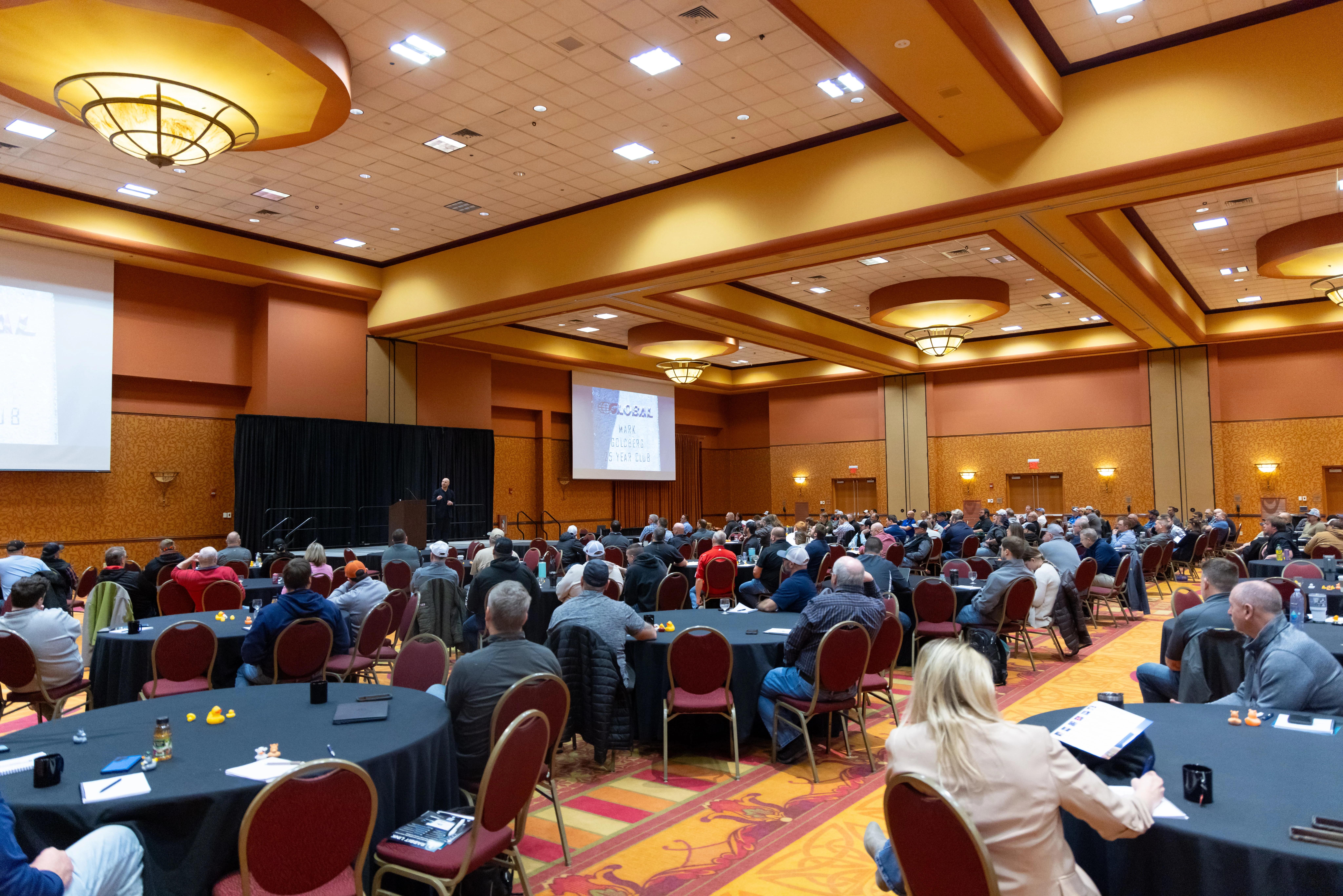 Attendees seated at round tables in a ballroom watching Danny Goldberg speak