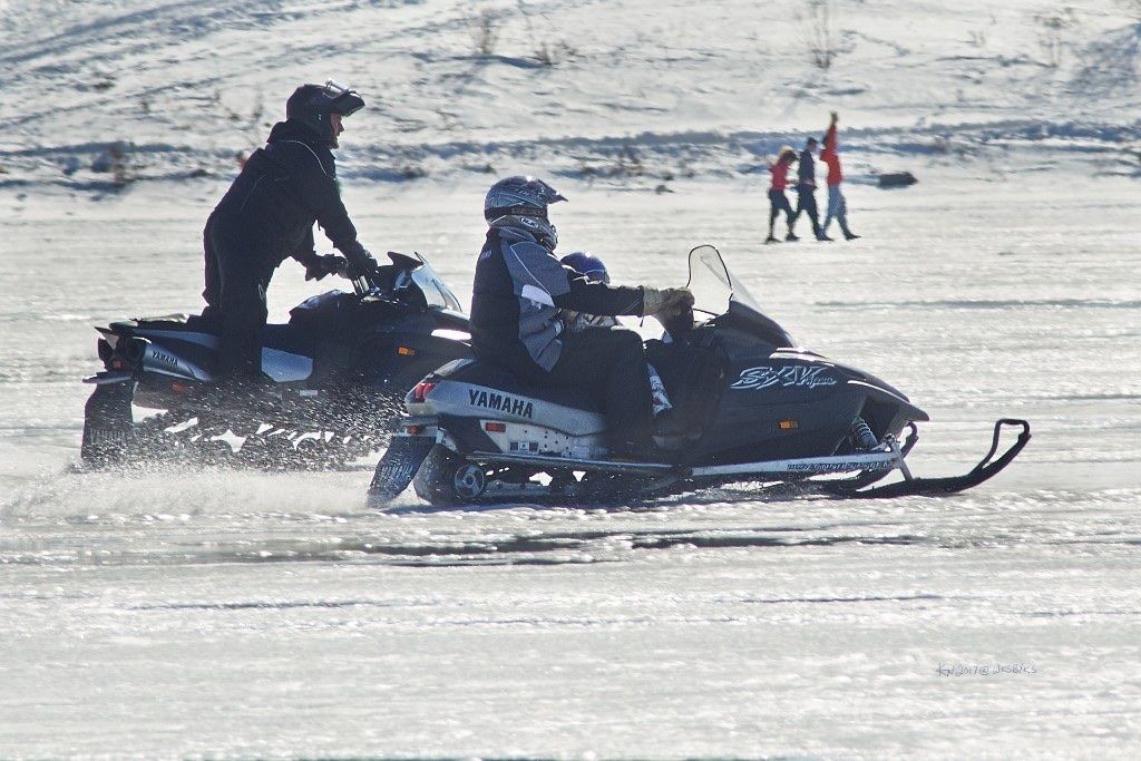 Two Yamaha snowmobiles racing across open ice with a child riding up front