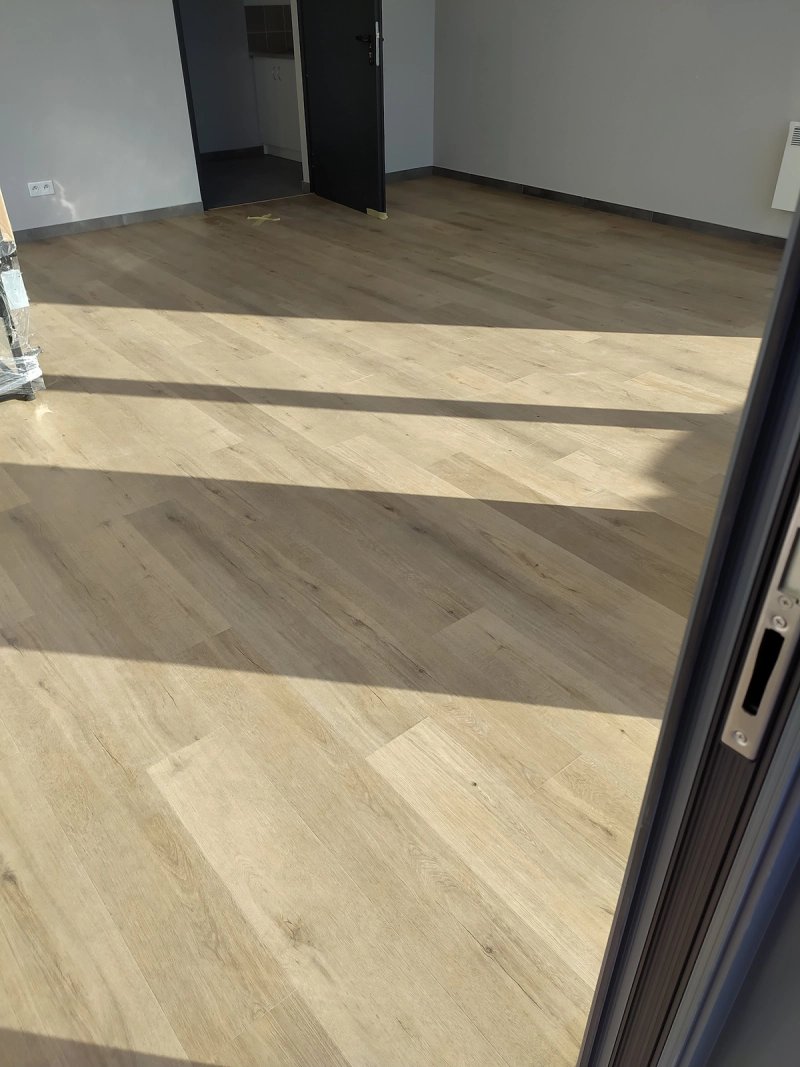 Pokój z nowo ułożonymi panelami podłogowymi w jasnym odcieniu dębu, promienie słońca przez okno rzucają cienie na podłodze