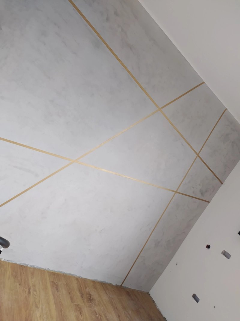 Detal ściany z tynkiem dekoracyjnym: marmurowa faktura i złote linie tworzące geometryczny wzór