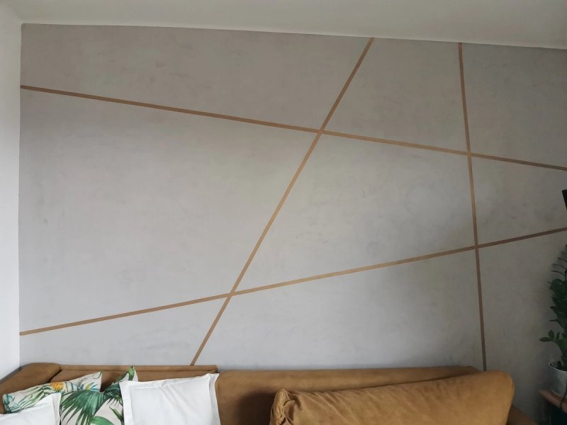 Salon z tynkiem dekoracyjnym w jasnym kolorze z dekoracyjnymi geometrycznymi złotymi liniami, karmelowa sofa i poduszki