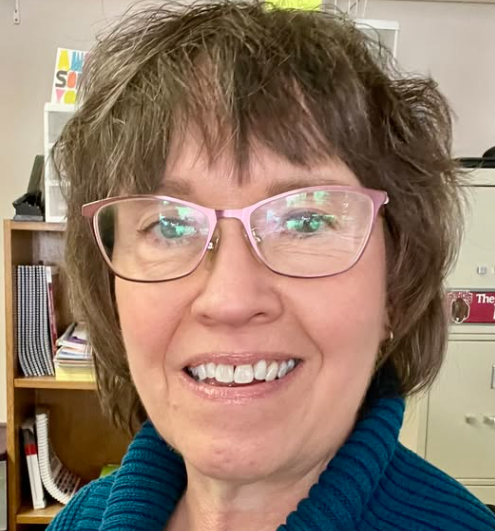 joanie.png A woman with glasses smiling indoors, with books and shelves in the background.