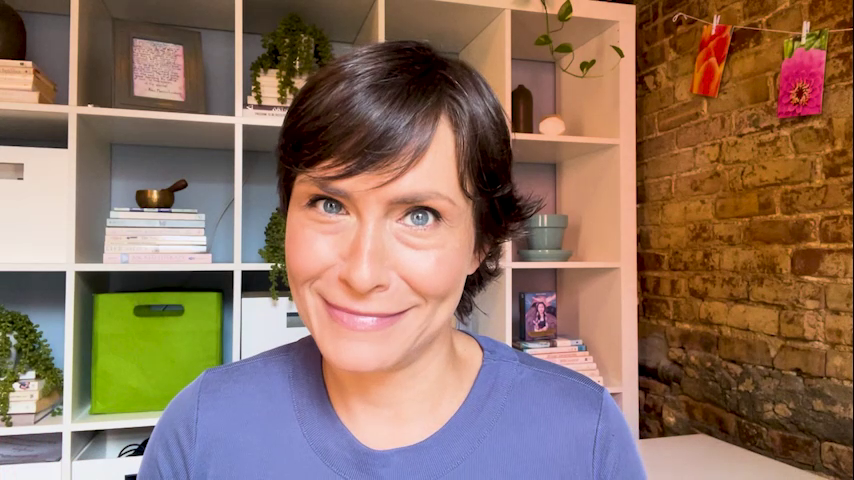 Person with short brown hair smiling in front of white shelving and a brick wall, with plants, books, and decorative items on the shelves.