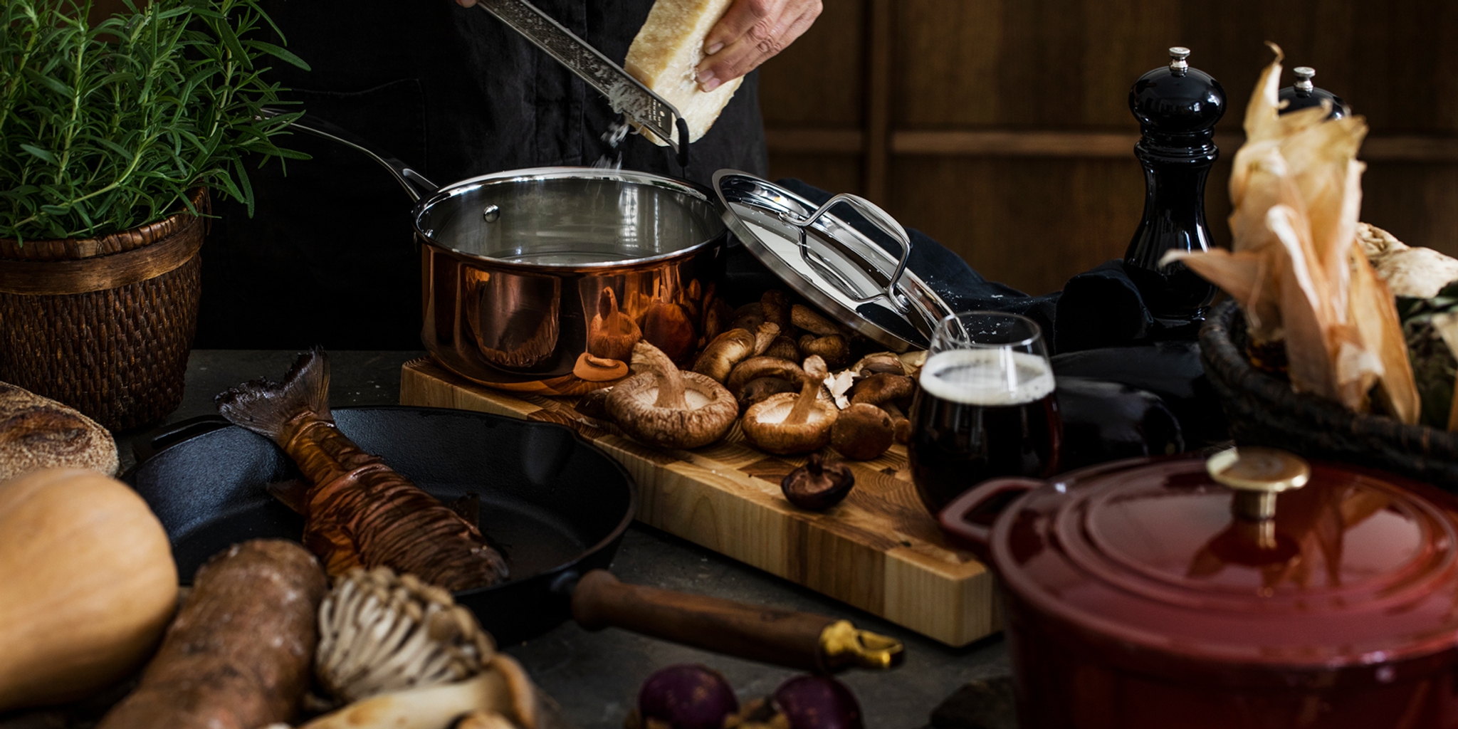 En person river ost i en kopparkastrull på ett rustikt bord fyllt med färska ingredienser som svamp, fisk och grönsaker.