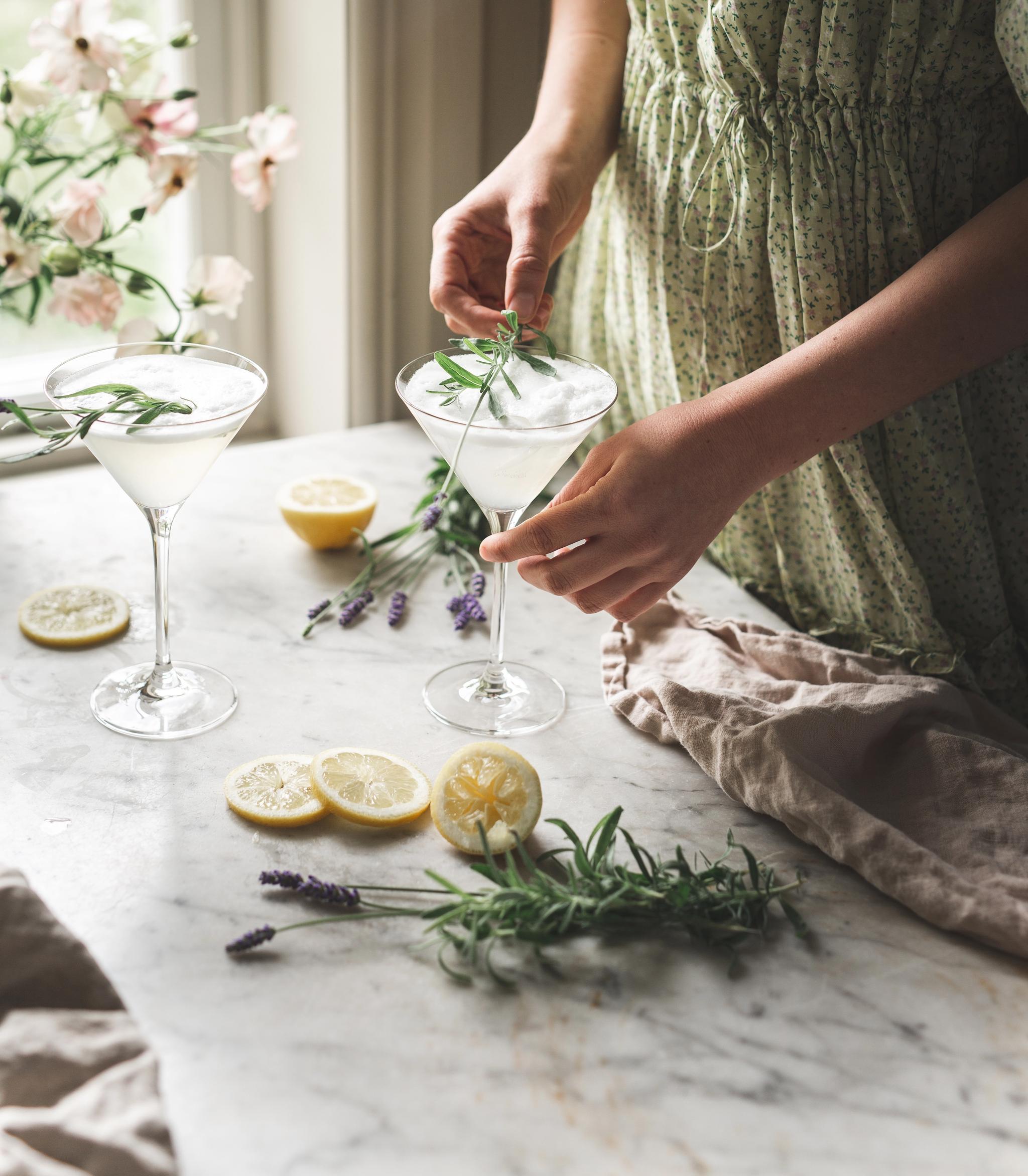 Kvinna i ljusgrön klänning dekorerar två eleganta citron- och rosmarindrinkar vid ett solbelyst köksfönster.
