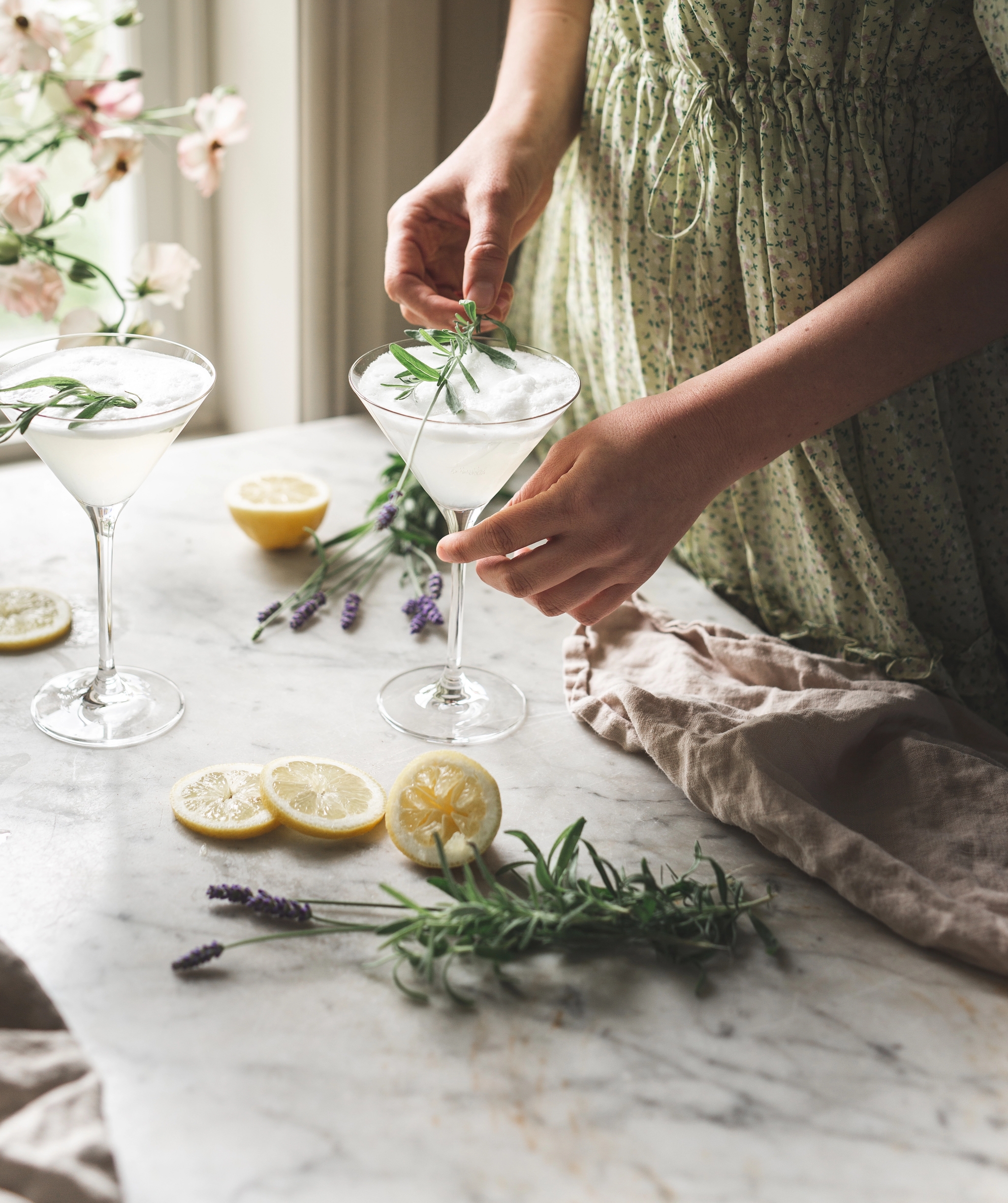 Kvinna i ljusgrön klänning dekorerar två somriga cocktails med rosmarin och citron på ett marmorbord vid ett ljust fönster.