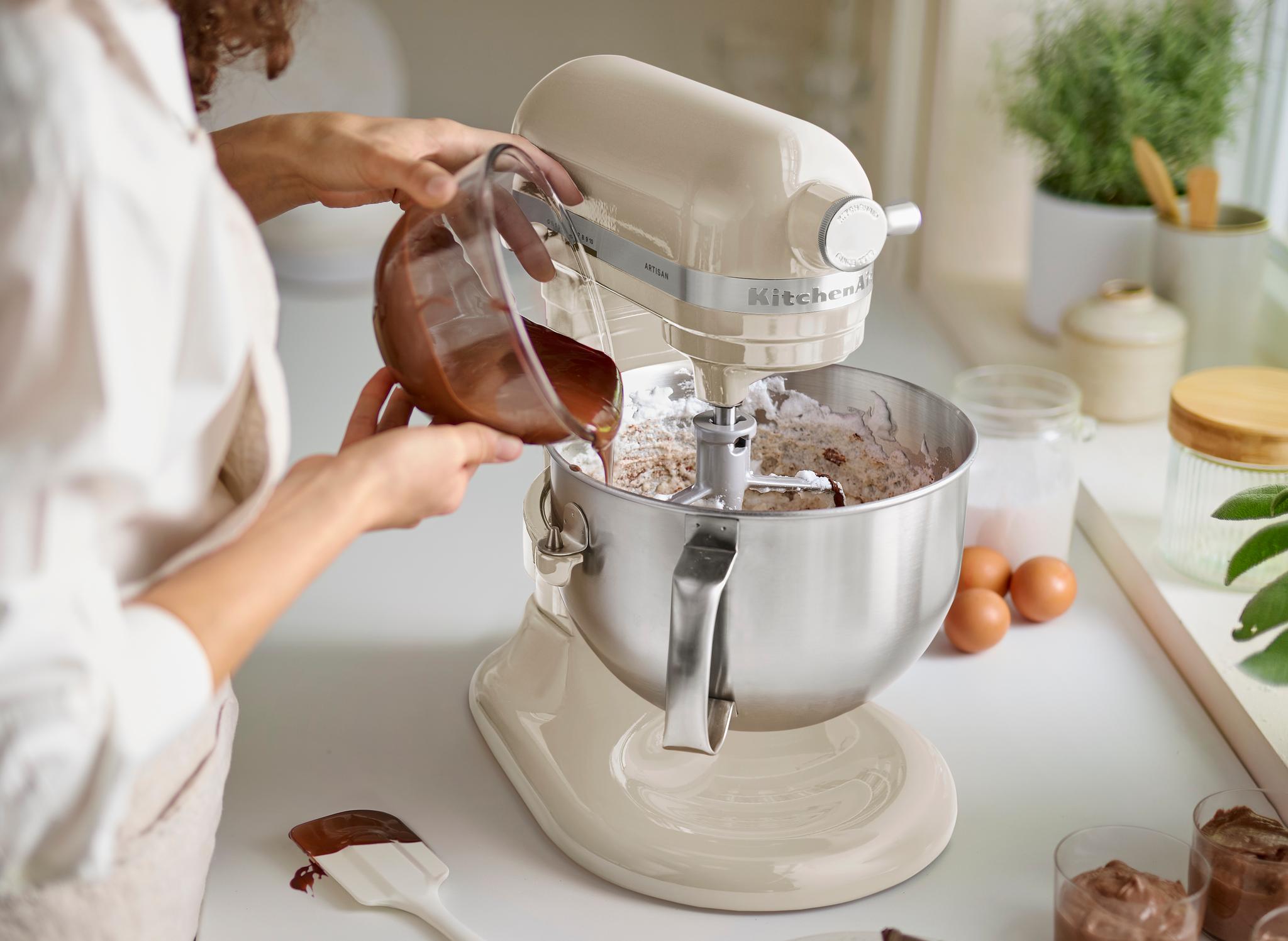 En person heller smeltet sjokolade fra et målebeger over i en kremfarget KitchenAid-standmikserbolle med tørre ingredienser.