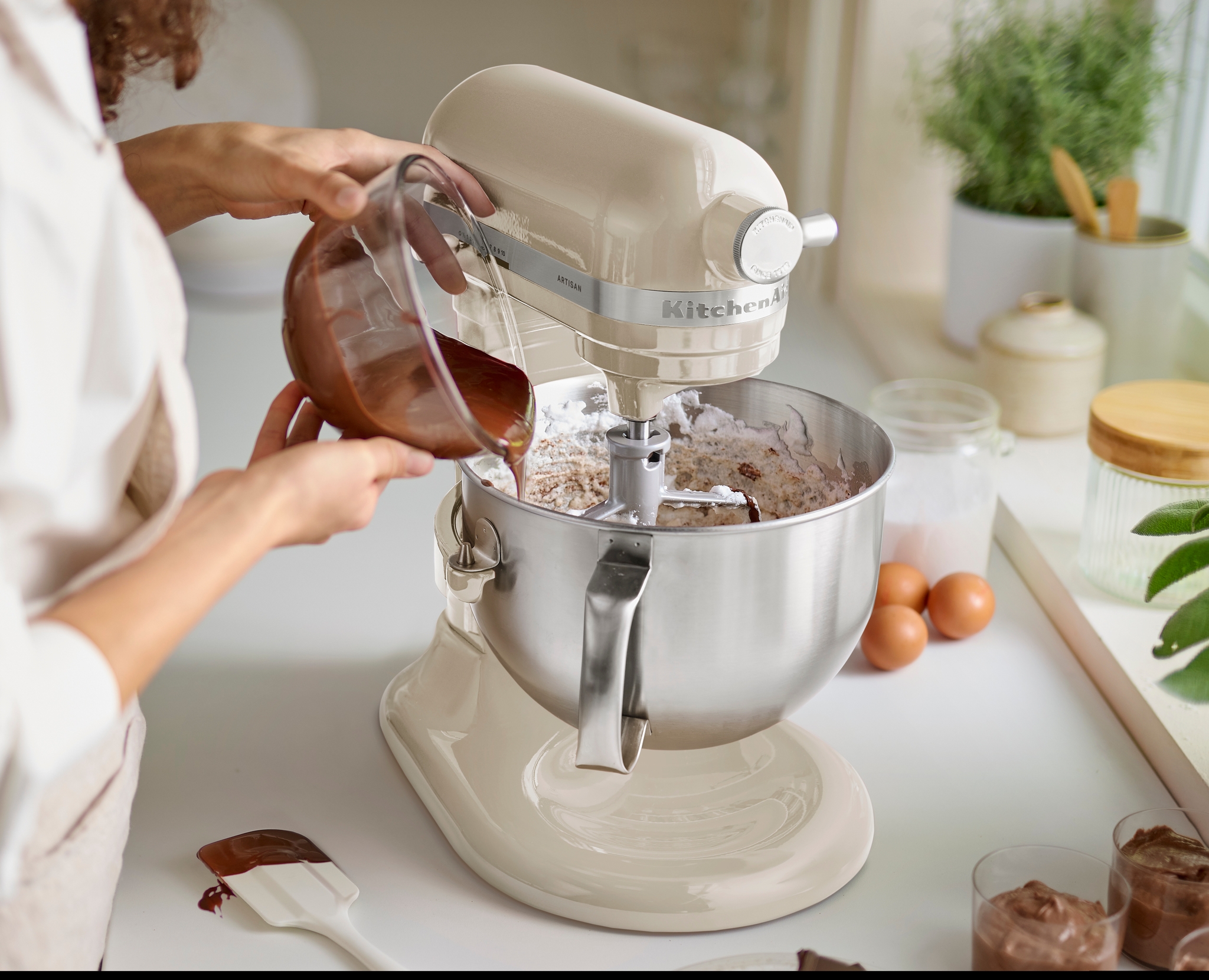 En person heller smeltet sjokolade fra et målebeger over i en kremfarget KitchenAid-standmikserbolle med tørre ingredienser.